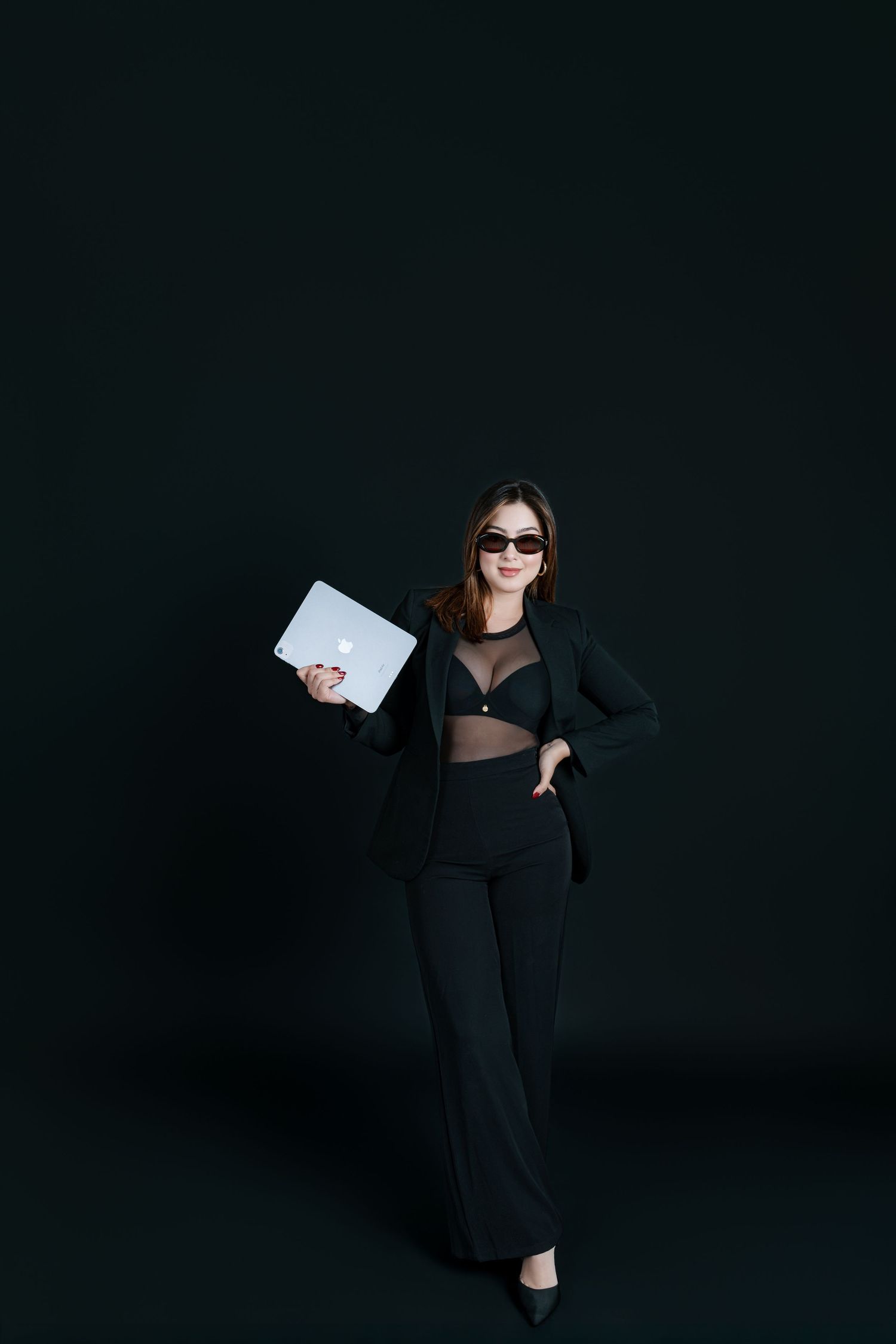 Stylish woman in black suit holding laptop wearing sunglasses against dark background.