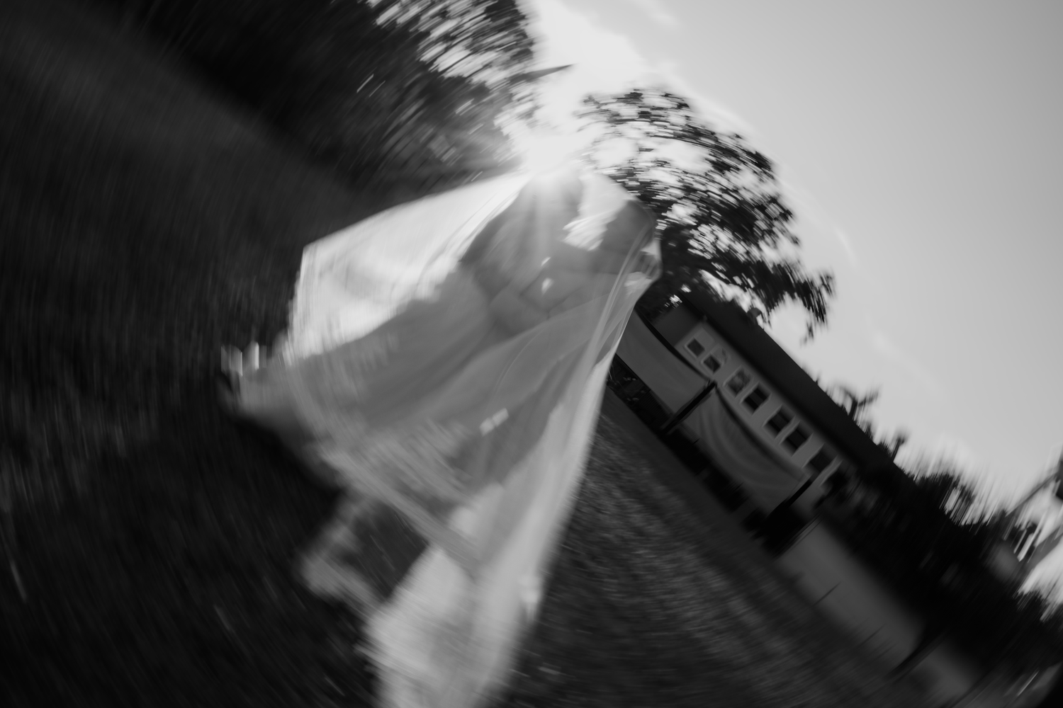 Black and white photo of a blurred wedding veil flowing near a vintage train and trees in soft sunlight.
