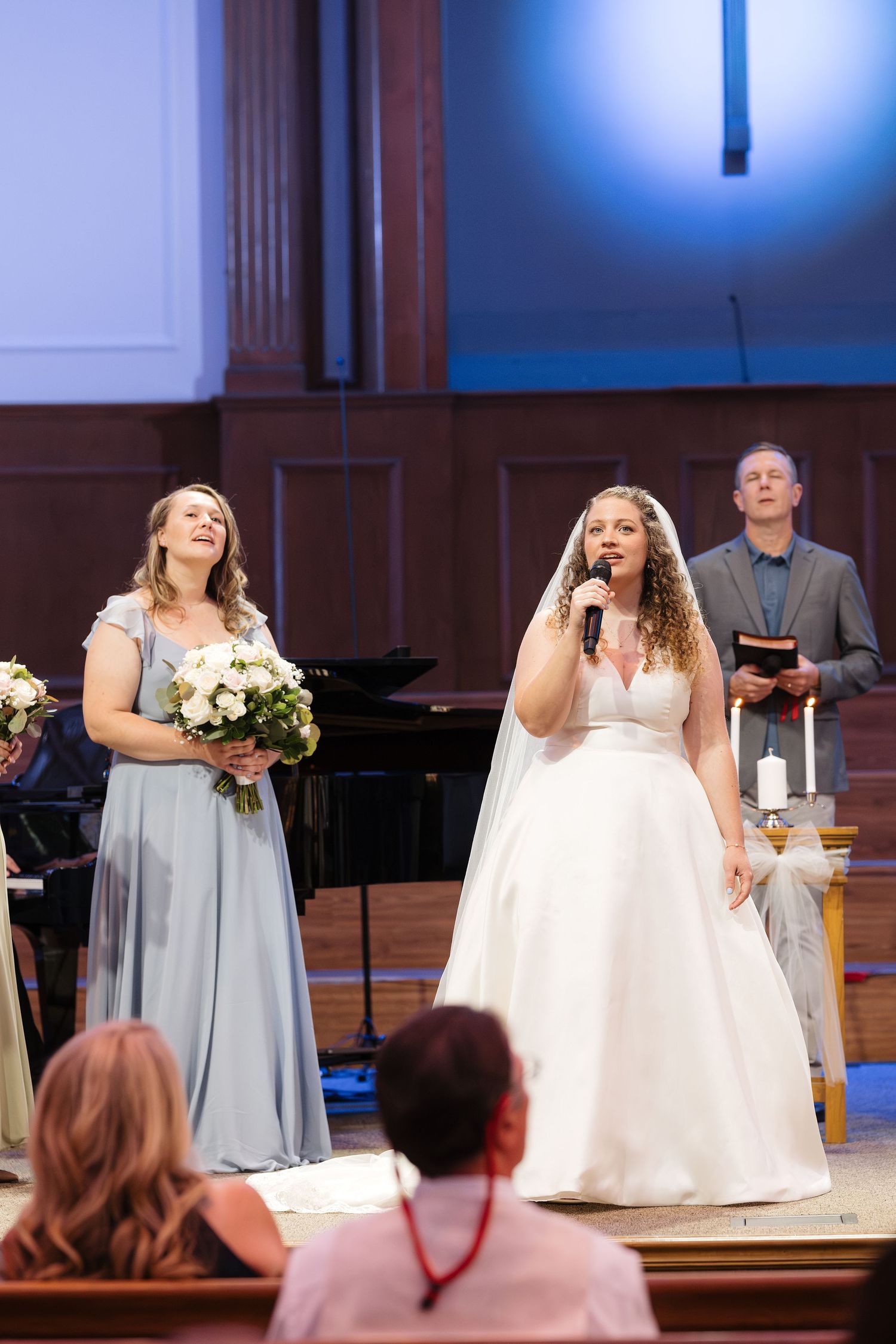 Bride singing into microphone at church altar in white gown beside bridesmaid in blue dress during wedding ceremony.