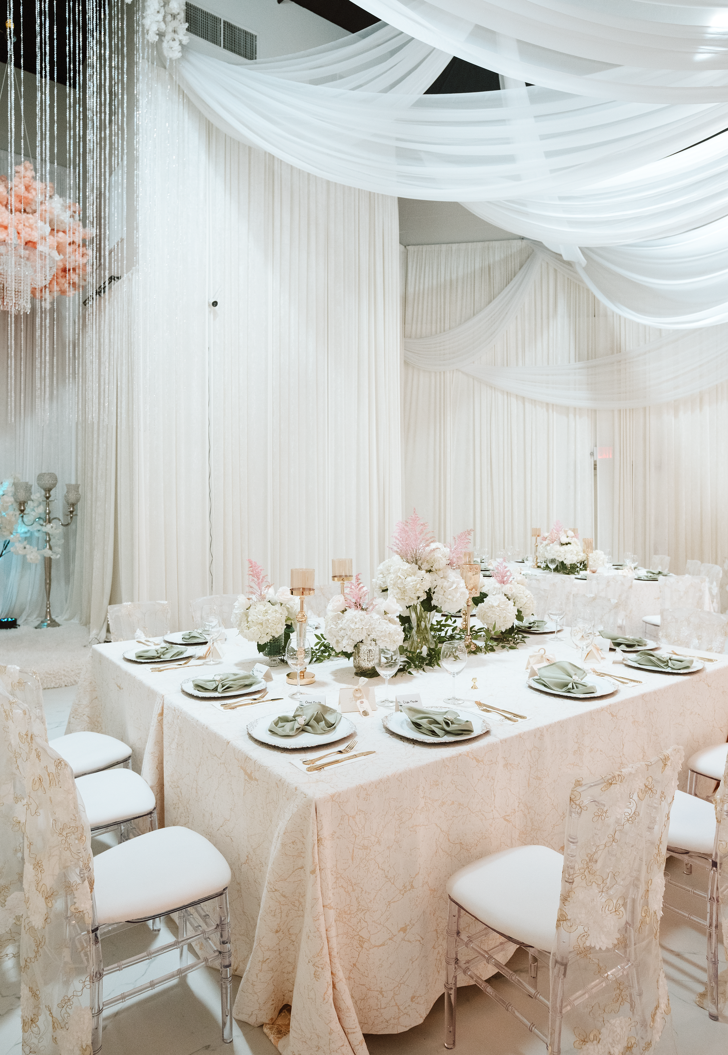 Elegant white wedding reception table with floral centerpieces, draped fabric backdrop, and acrylic chairs in a luxurious venue.
