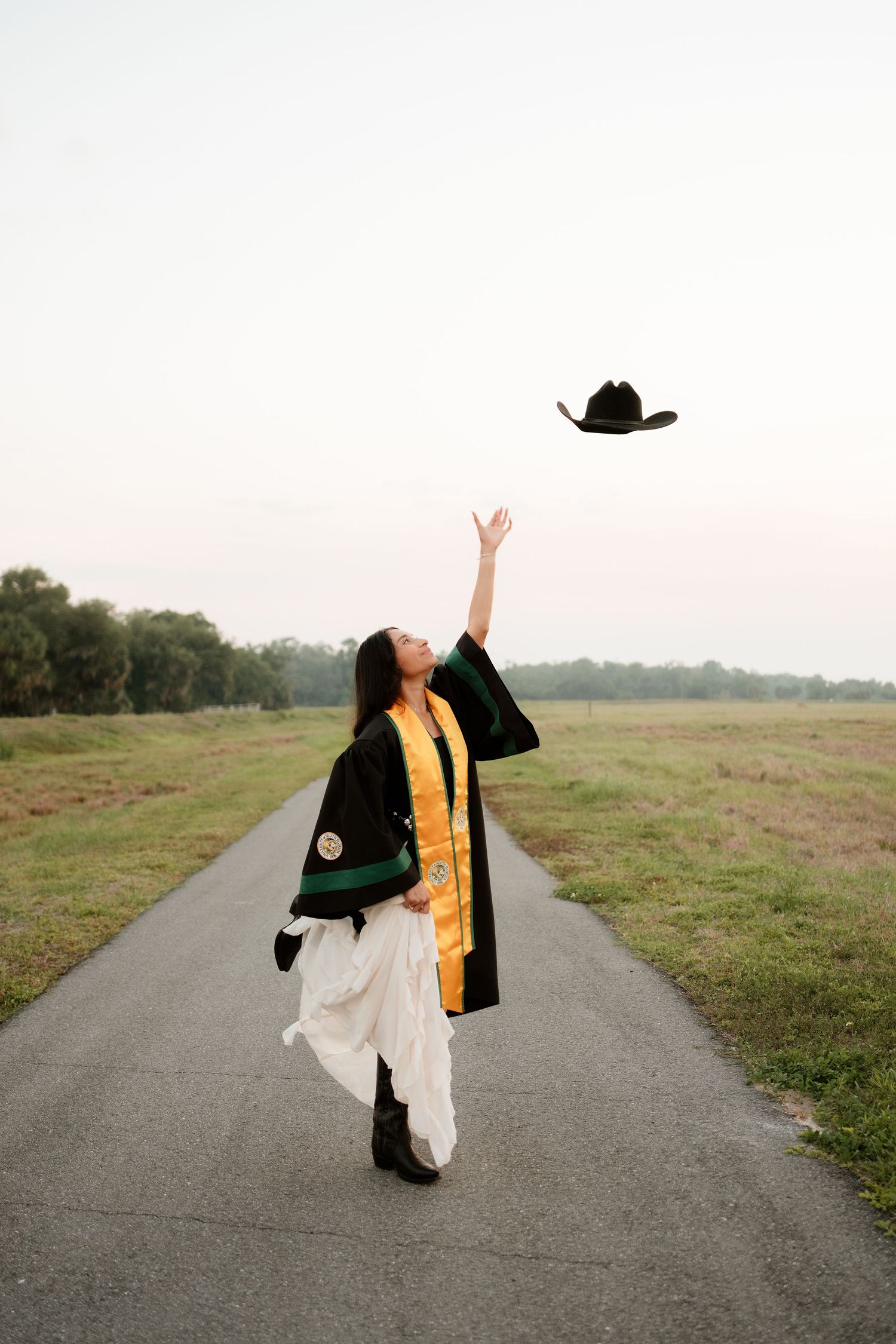 Graduate in black robe and gold sash tosses cowboy hat in air on scenic path.