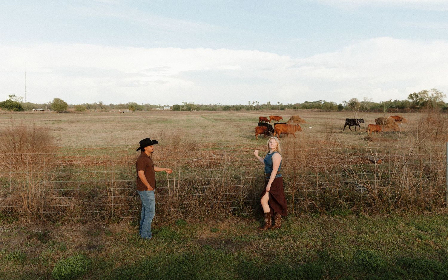 Two people stand by a fence on a rural Texas ranch with cattle grazing in a dry field behind them.