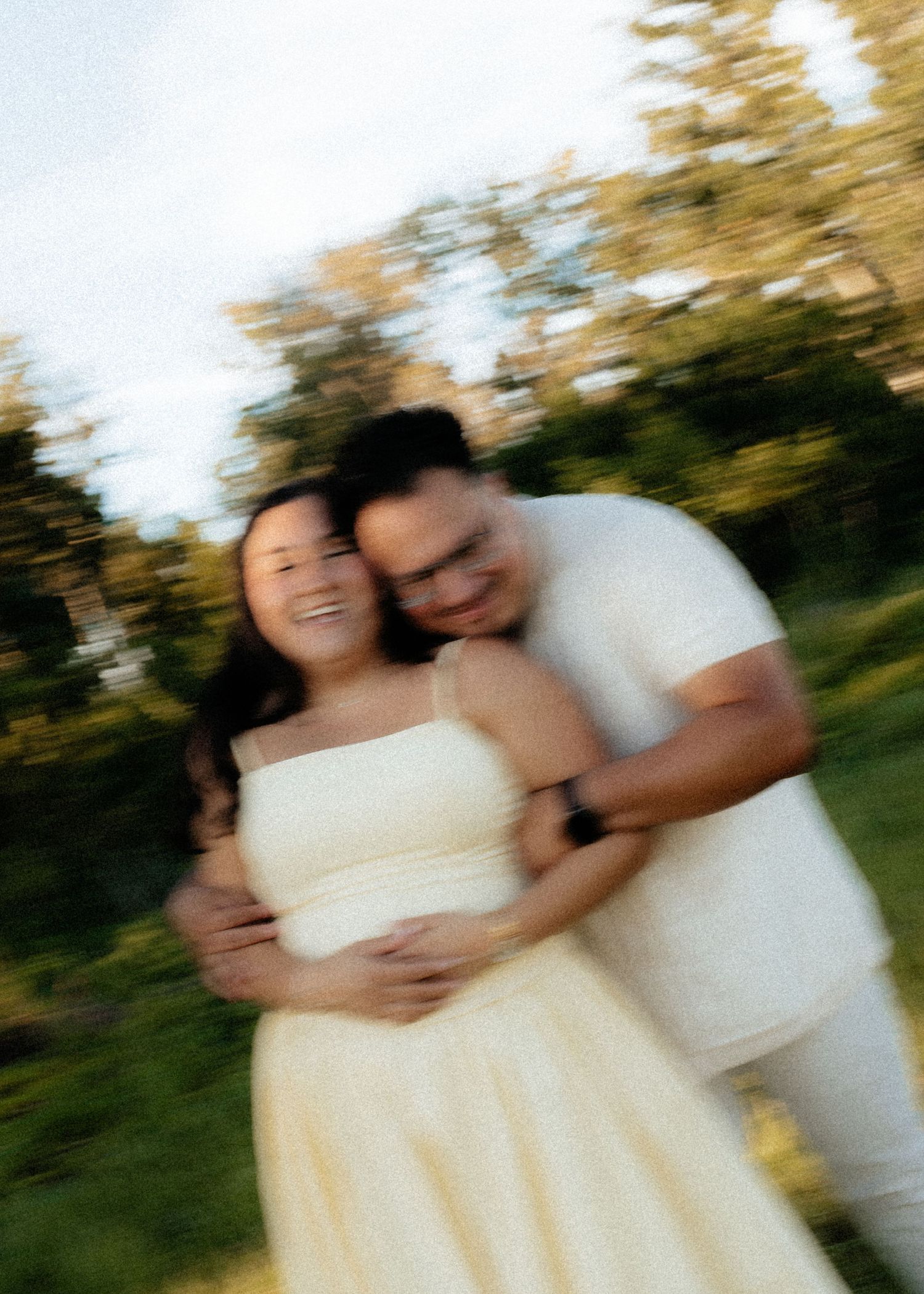 A smiling couple embraces outdoors, the woman in a yellow dress with the man in white behind her, in a dreamy blurred photo.