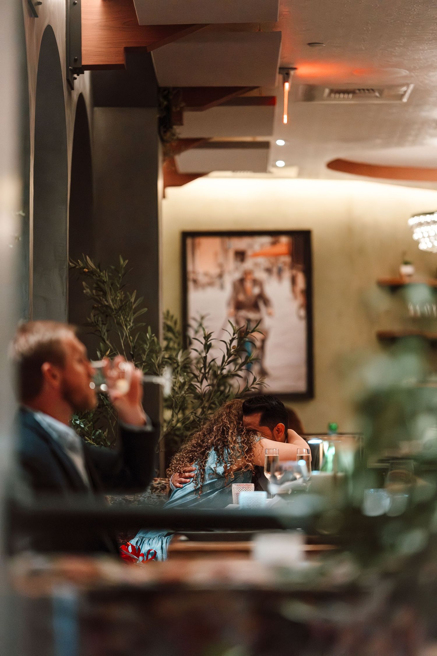 Cozy restaurant interior with patrons dining, lush greenery, warm lighting, and framed artwork on the wall.