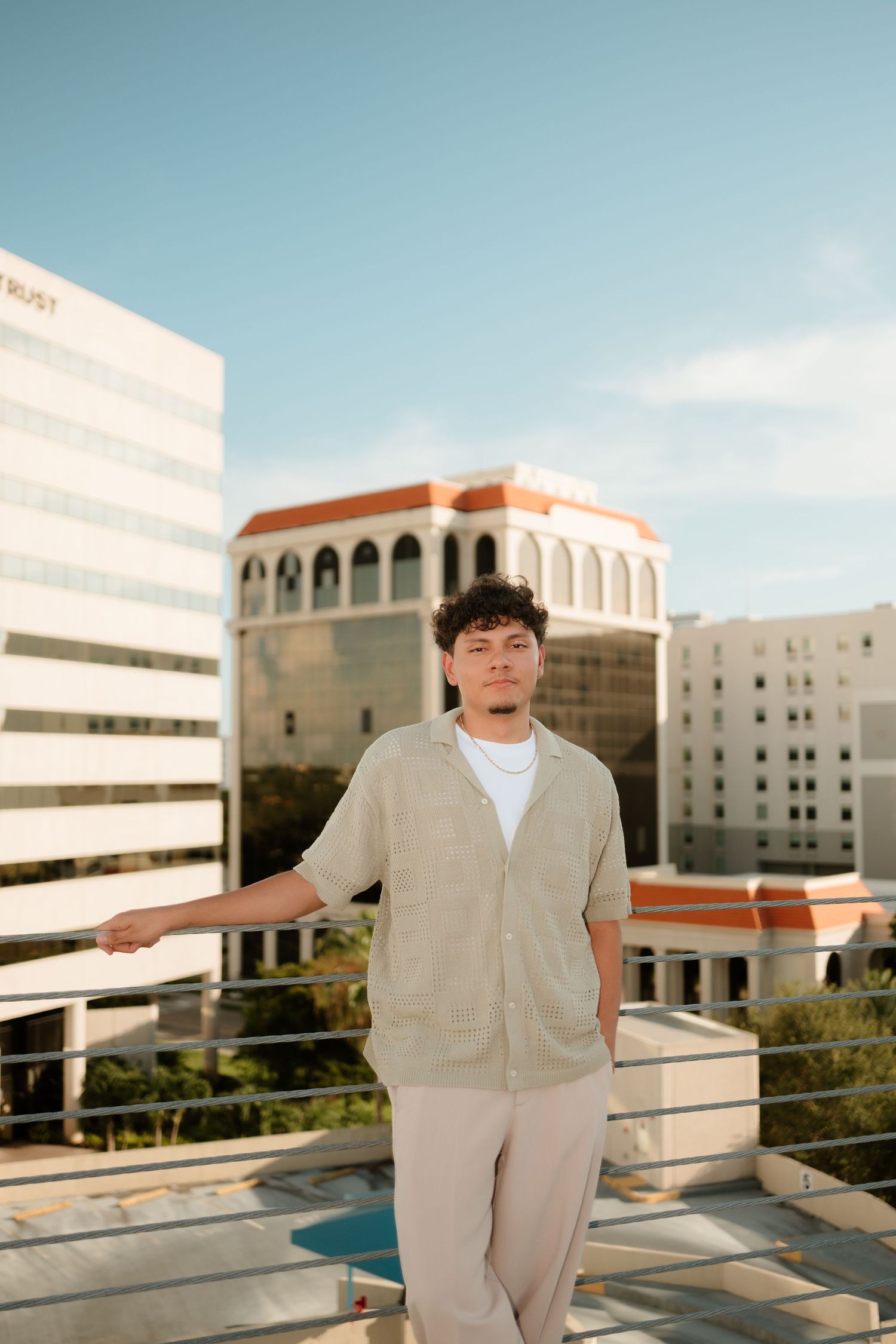 Young man in beige outfit poses on rooftop with city buildings and blue sky backdrop.