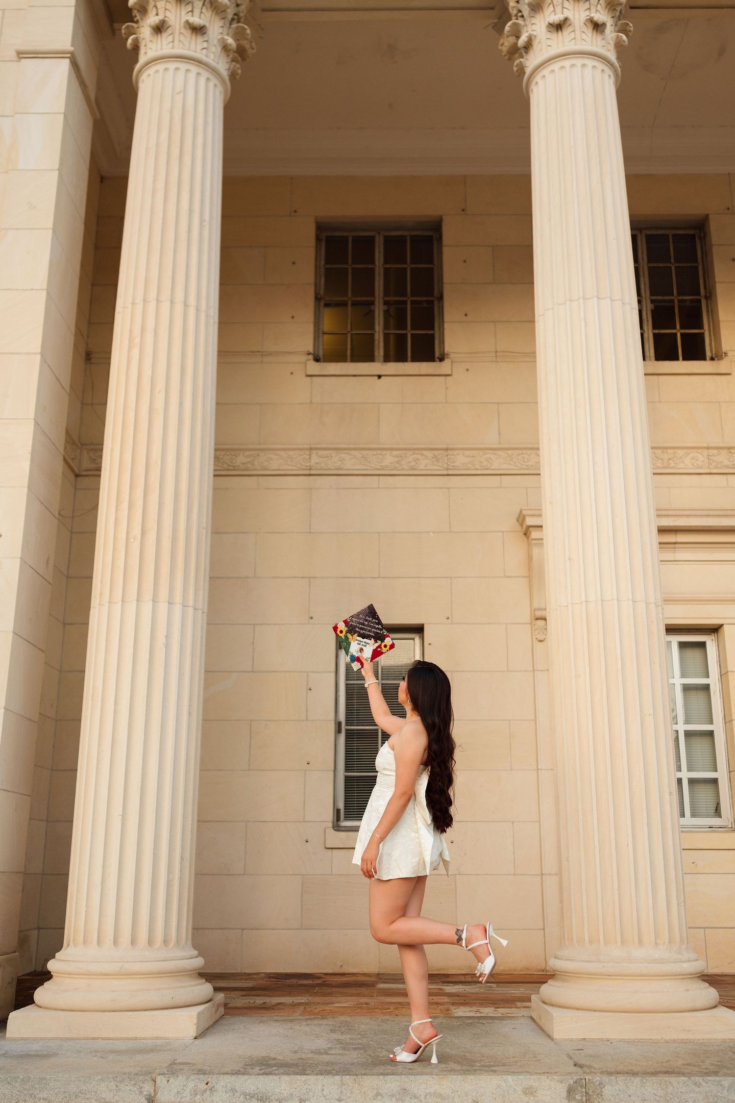 Young woman in white dress tossing graduation cap on steps of classical building with tall columns.