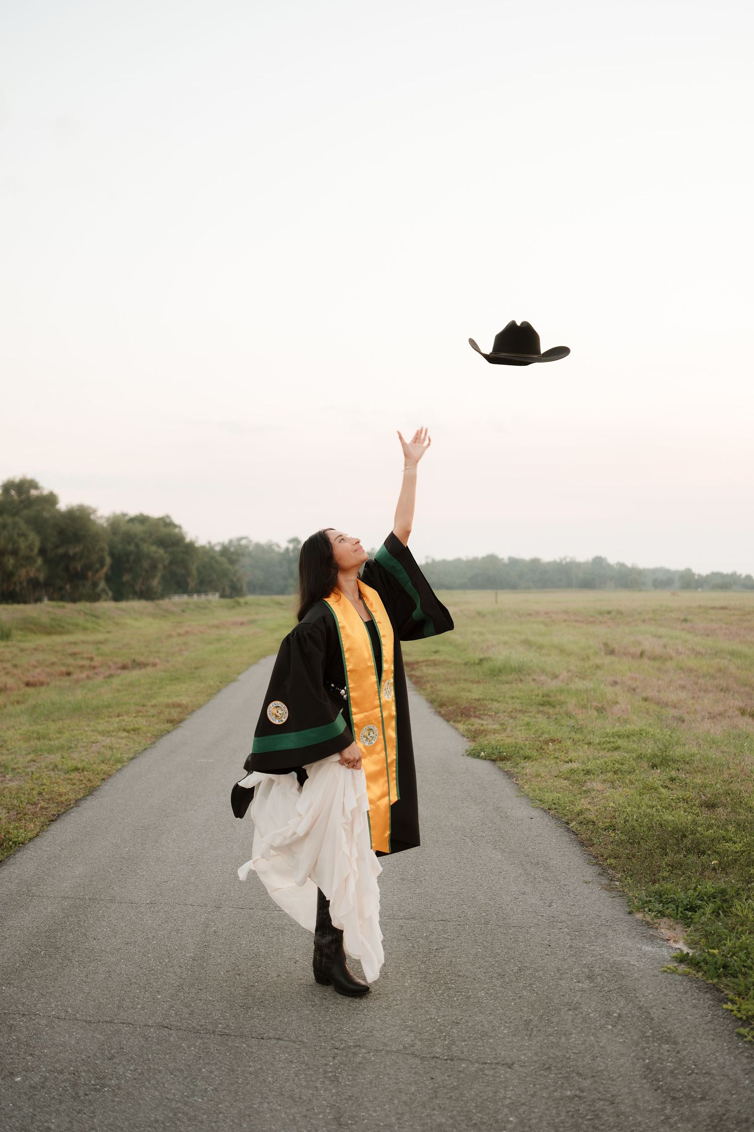 Graduate in black robe and gold stole tosses cowboy hat in air on scenic path.