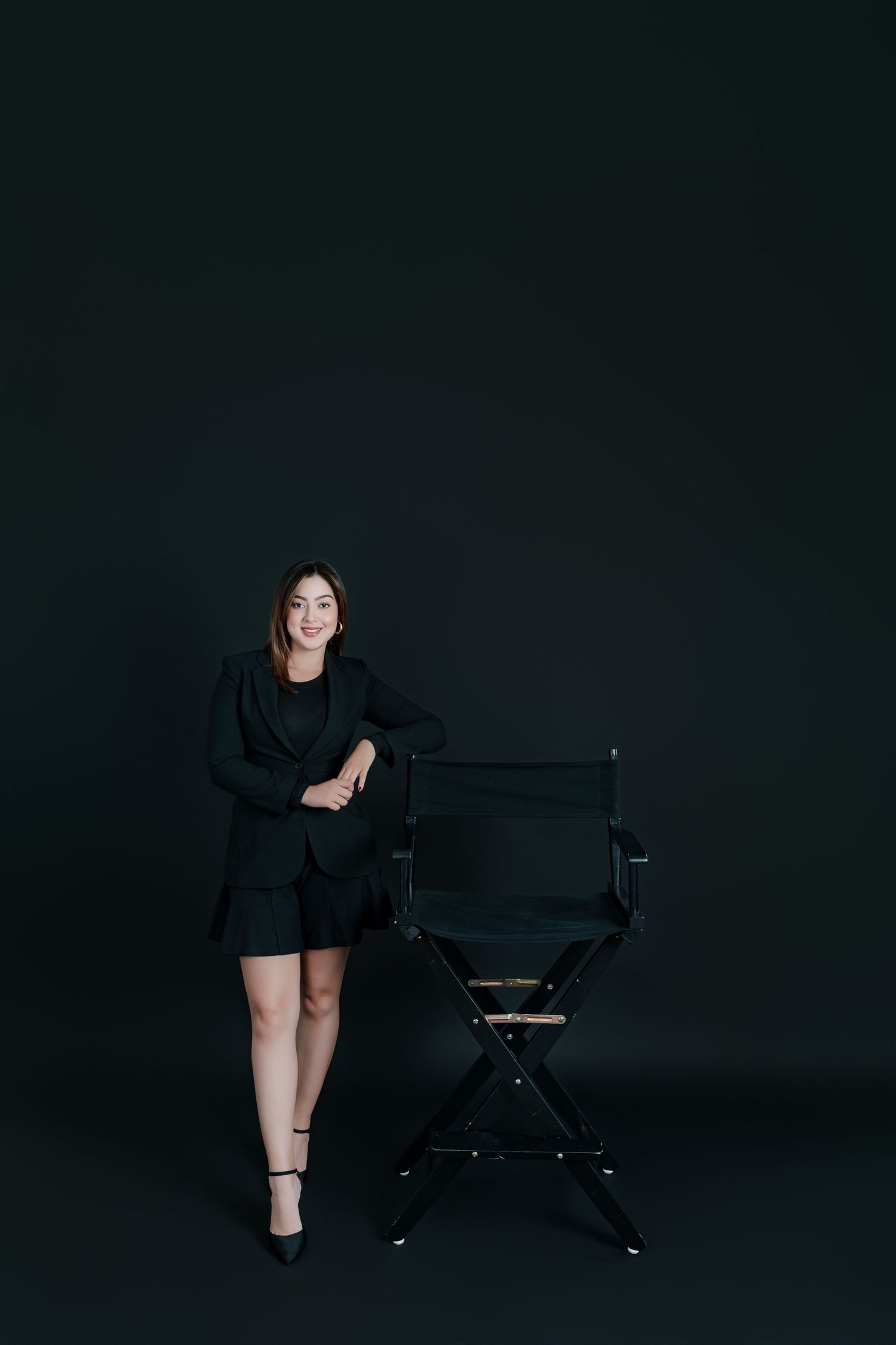 Woman in black blazer and shorts leaning on director's chair against dark background.