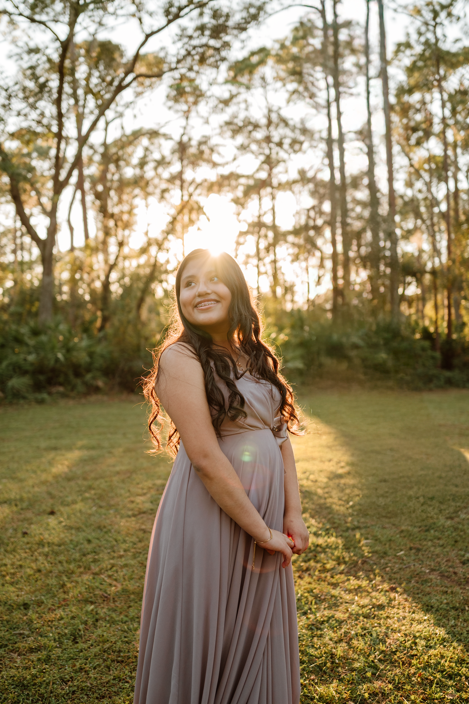 Smiling pregnant woman with long wavy hair in a flowing gray dress cradles her baby bump in a sunlit park at golden hour.