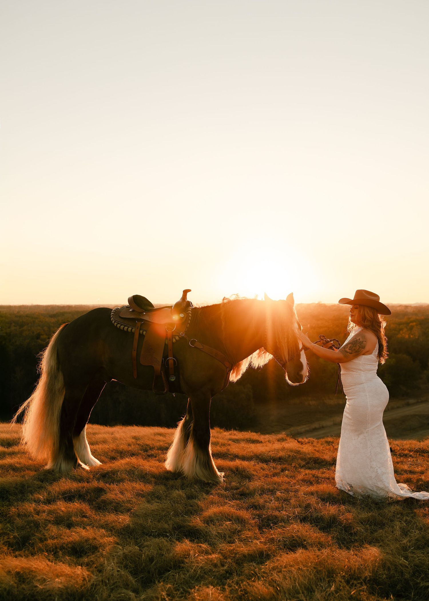 Bride in white gown and cowboy hat stands with a dark horse at golden sunset on a grassy field.