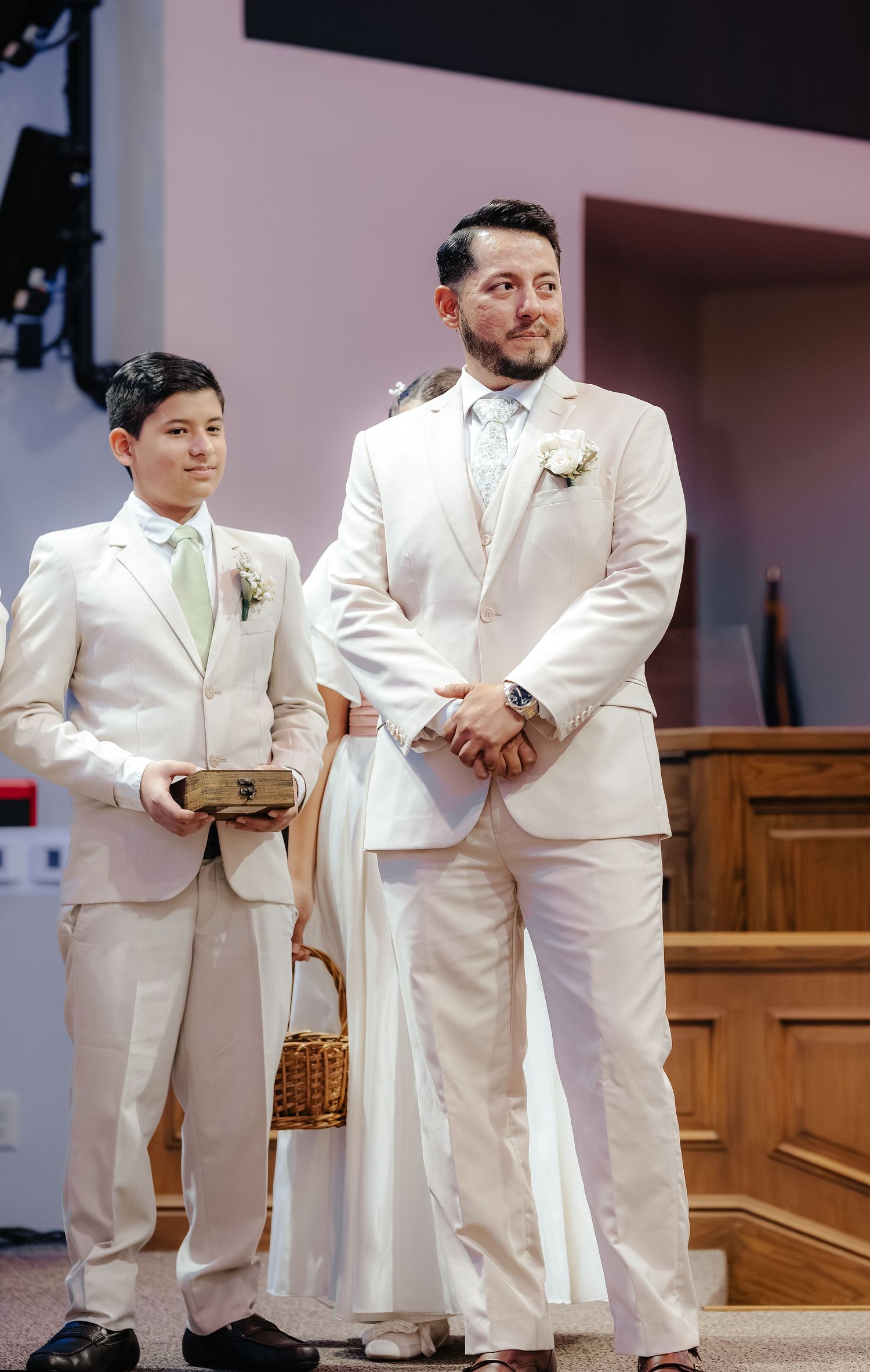 Groom and young ring bearer in matching white suits stand at the altar during a wedding ceremony in a church.