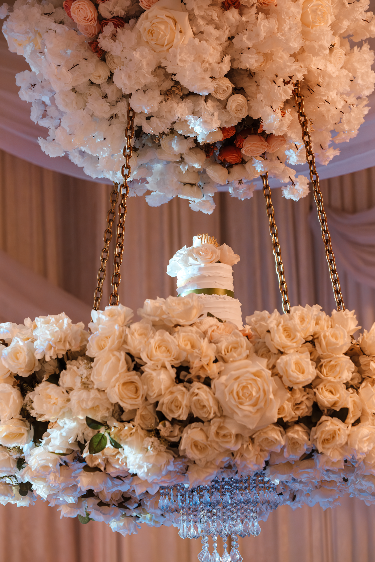 Stunning wedding cake display surrounded by lush white and blush roses on an ornate chandelier.