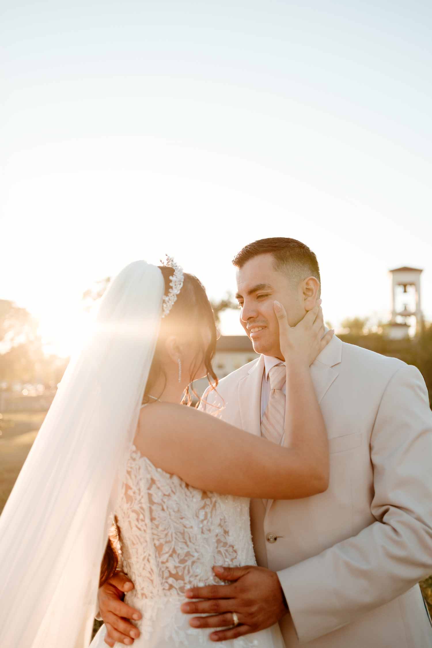 Bride and groom share a romantic golden hour moment, with bride touching groom's face in wedding attire.