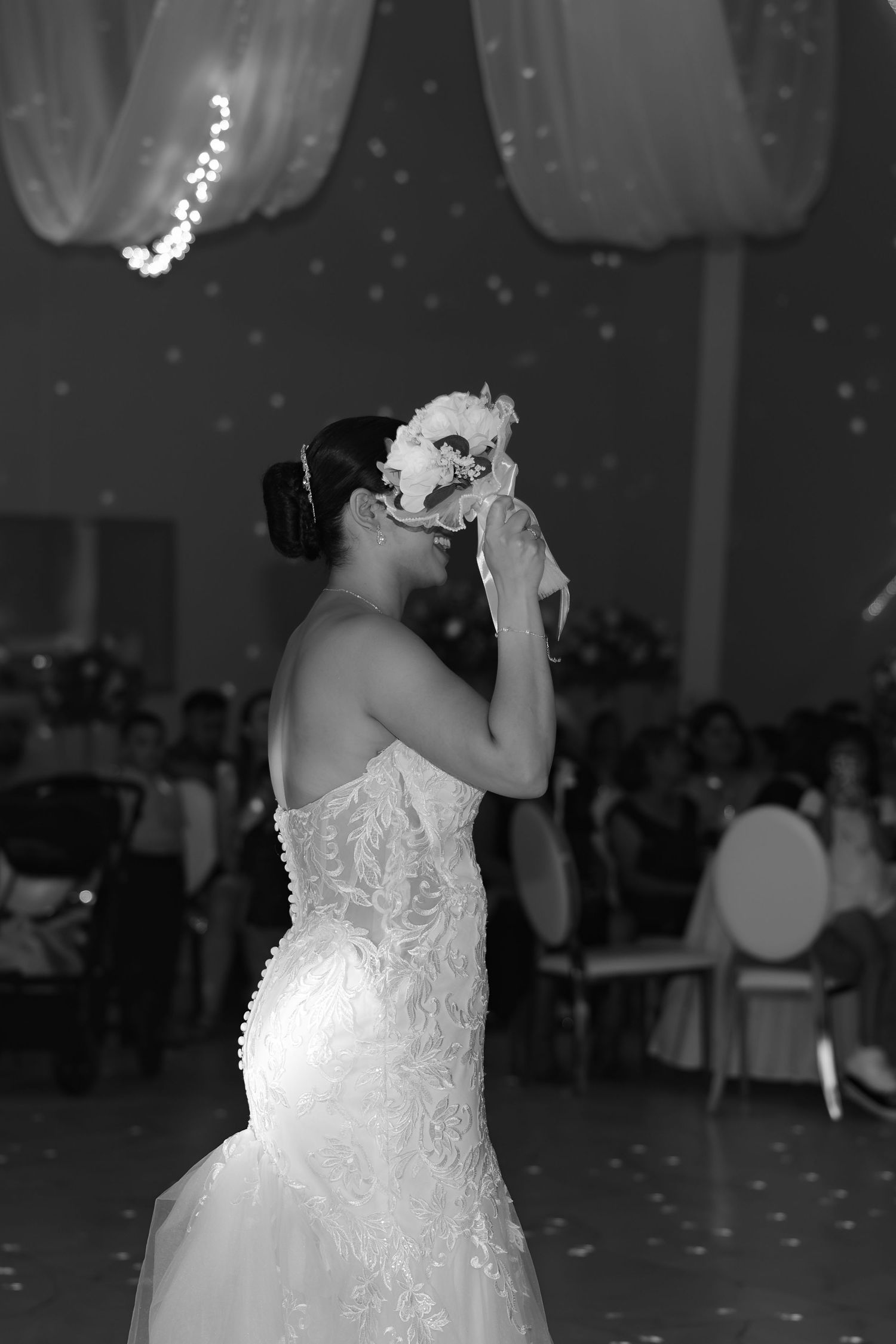 Black and white photo of a bride in lace gown holding bouquet to her face during wedding reception.