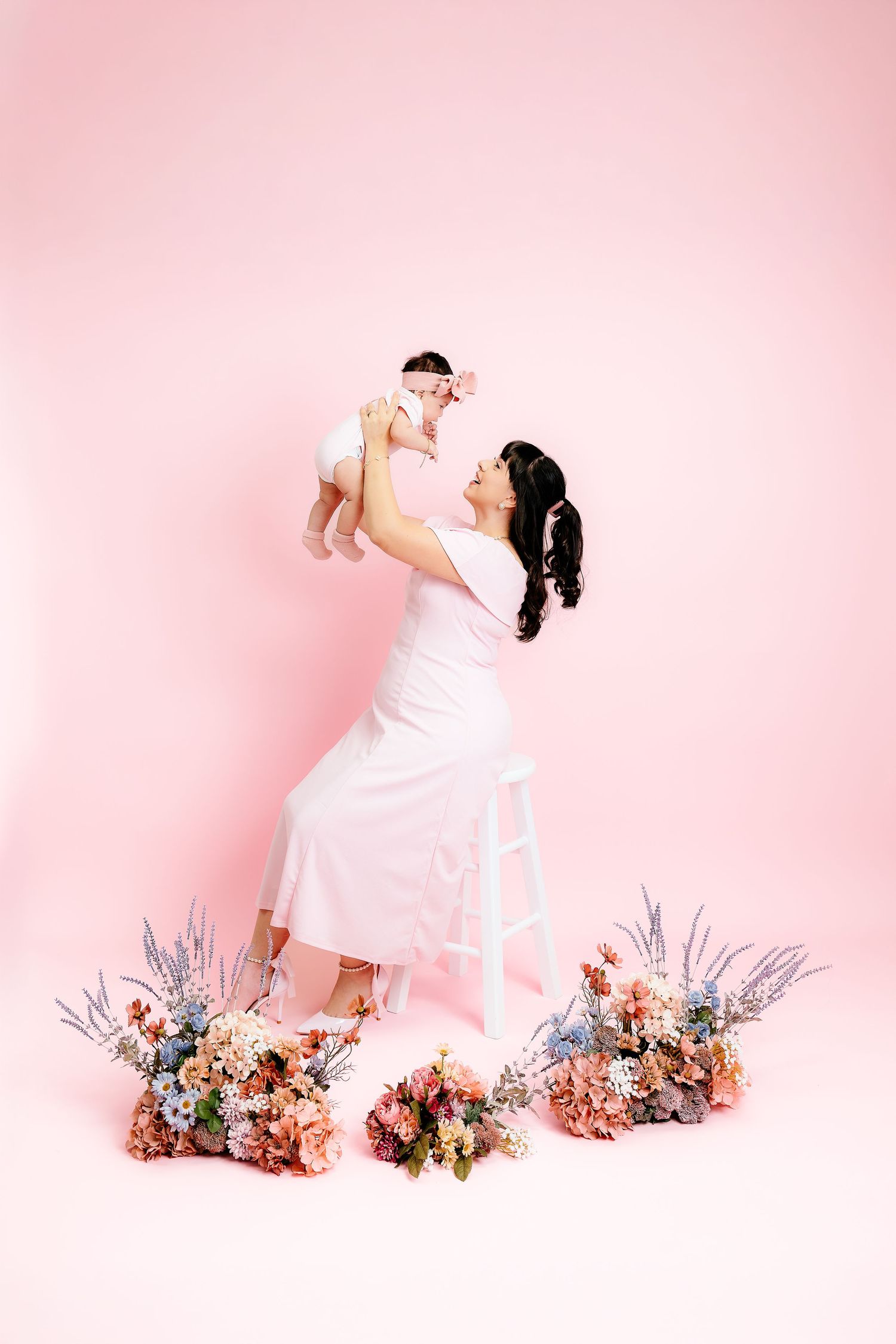 A mother in a white dress lifts her baby girl amid colorful flowers on a pink studio background.