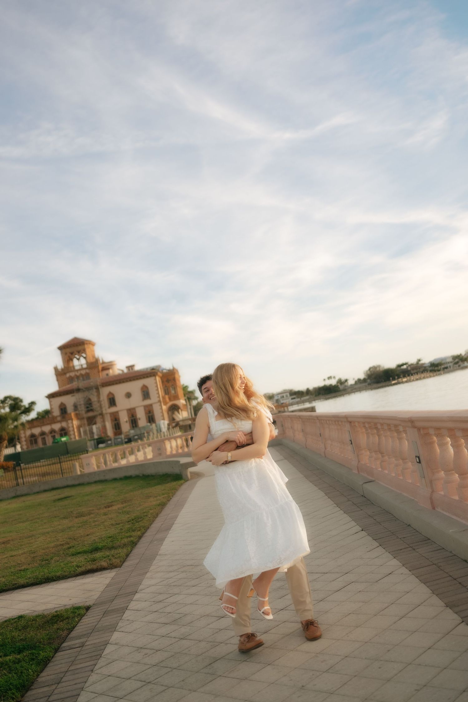 Couple dancing on waterfront promenade near historic building at golden hour, woman in white dress.