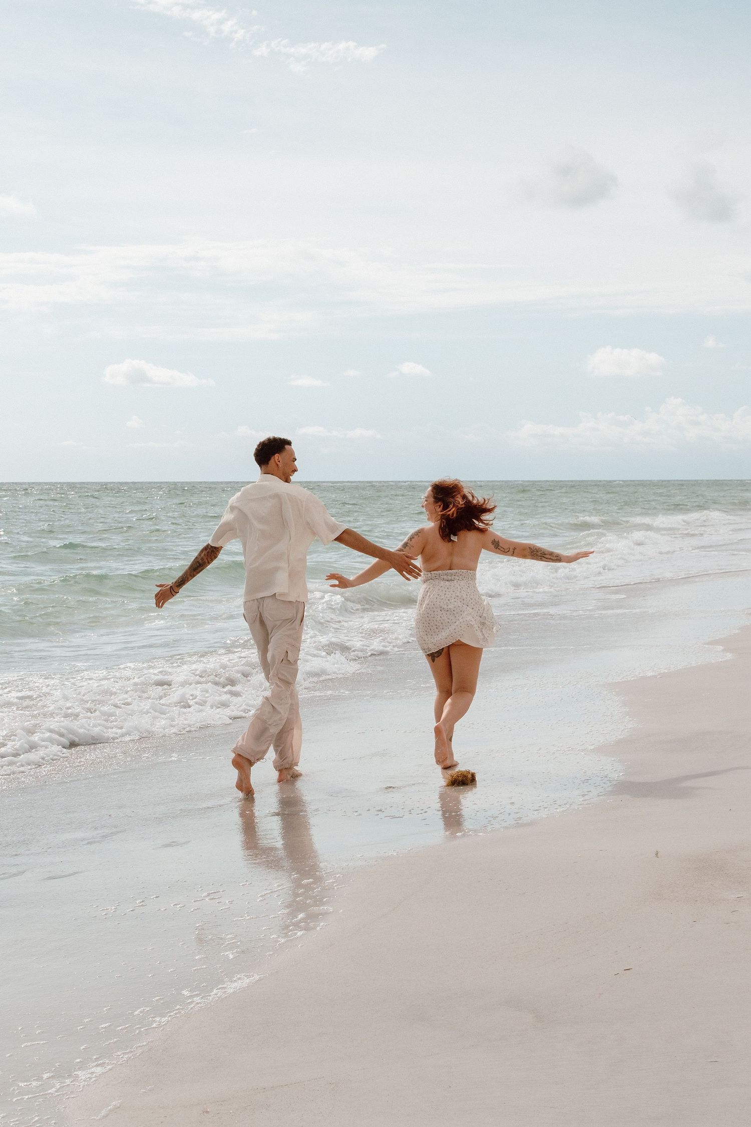 Couple holding hands running along a sandy beach shoreline in white outfits with ocean waves behind them.