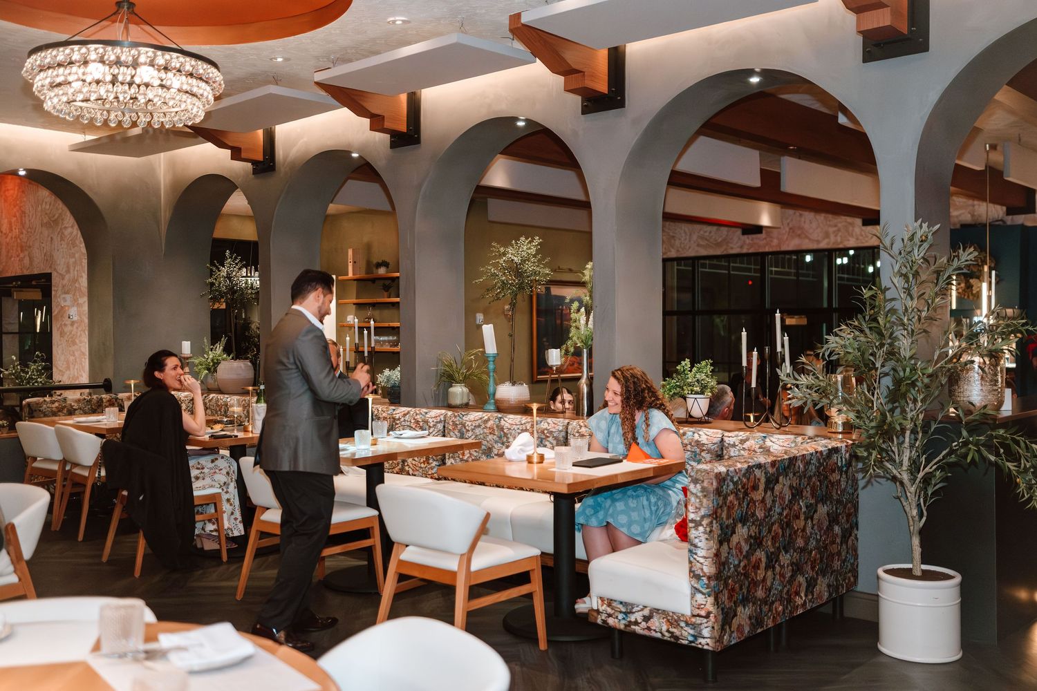 Elegant restaurant interior with arched walls, chandelier, floral booths, and a waiter serving guests at a candlelit table.