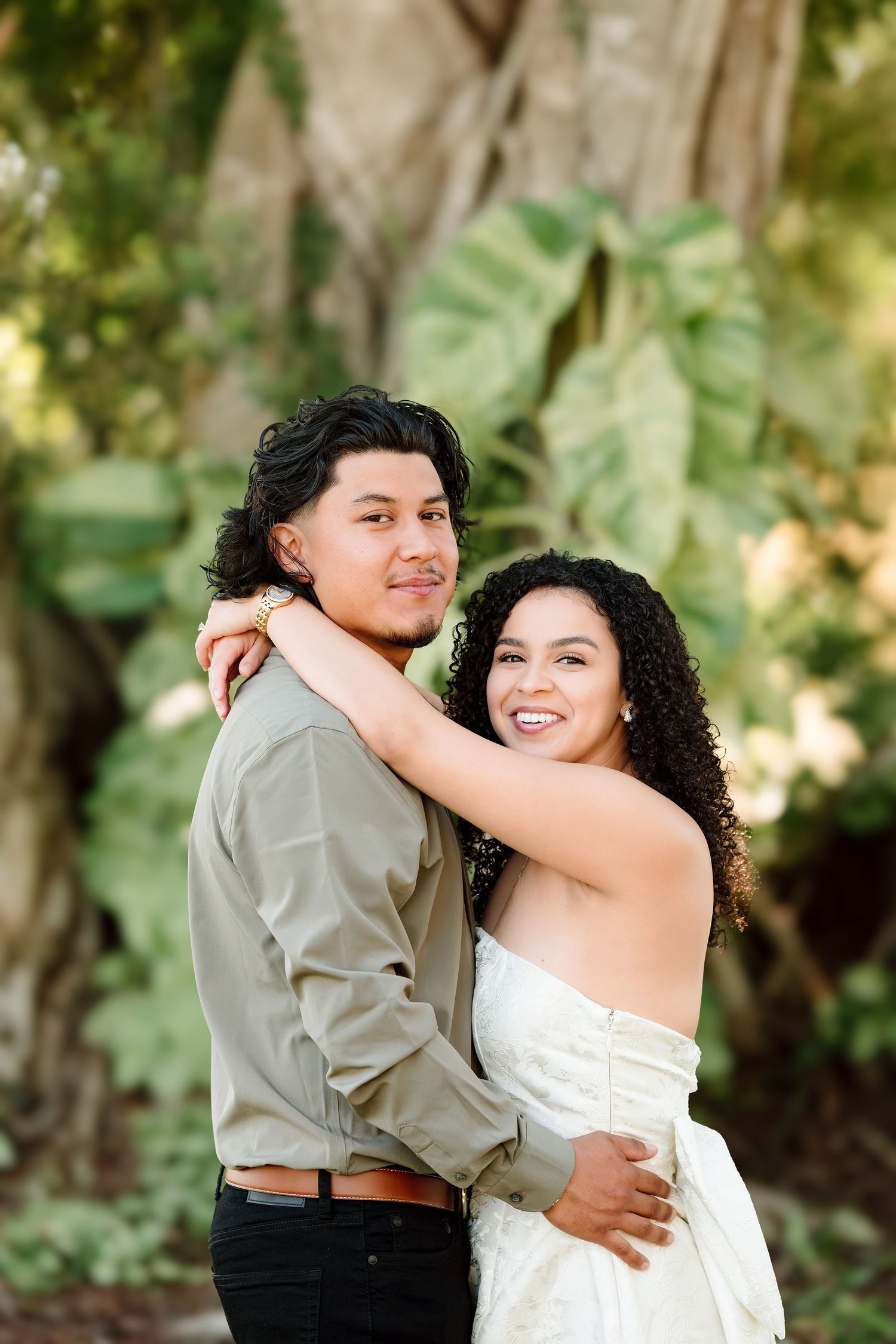 Couple embracing outdoors with lush green tropical foliage in background, woman in white dress smiling at camera.