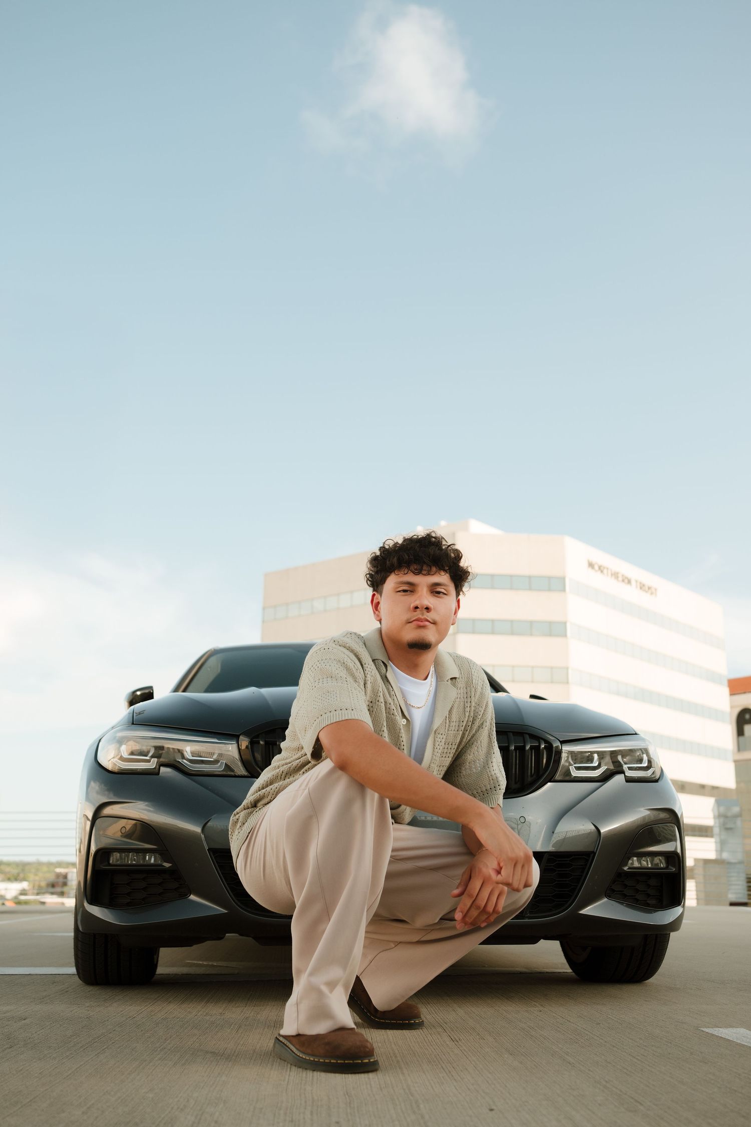 Young man crouching in front of a dark BMW sedan in a parking lot, wearing casual clothes with a building in the background.