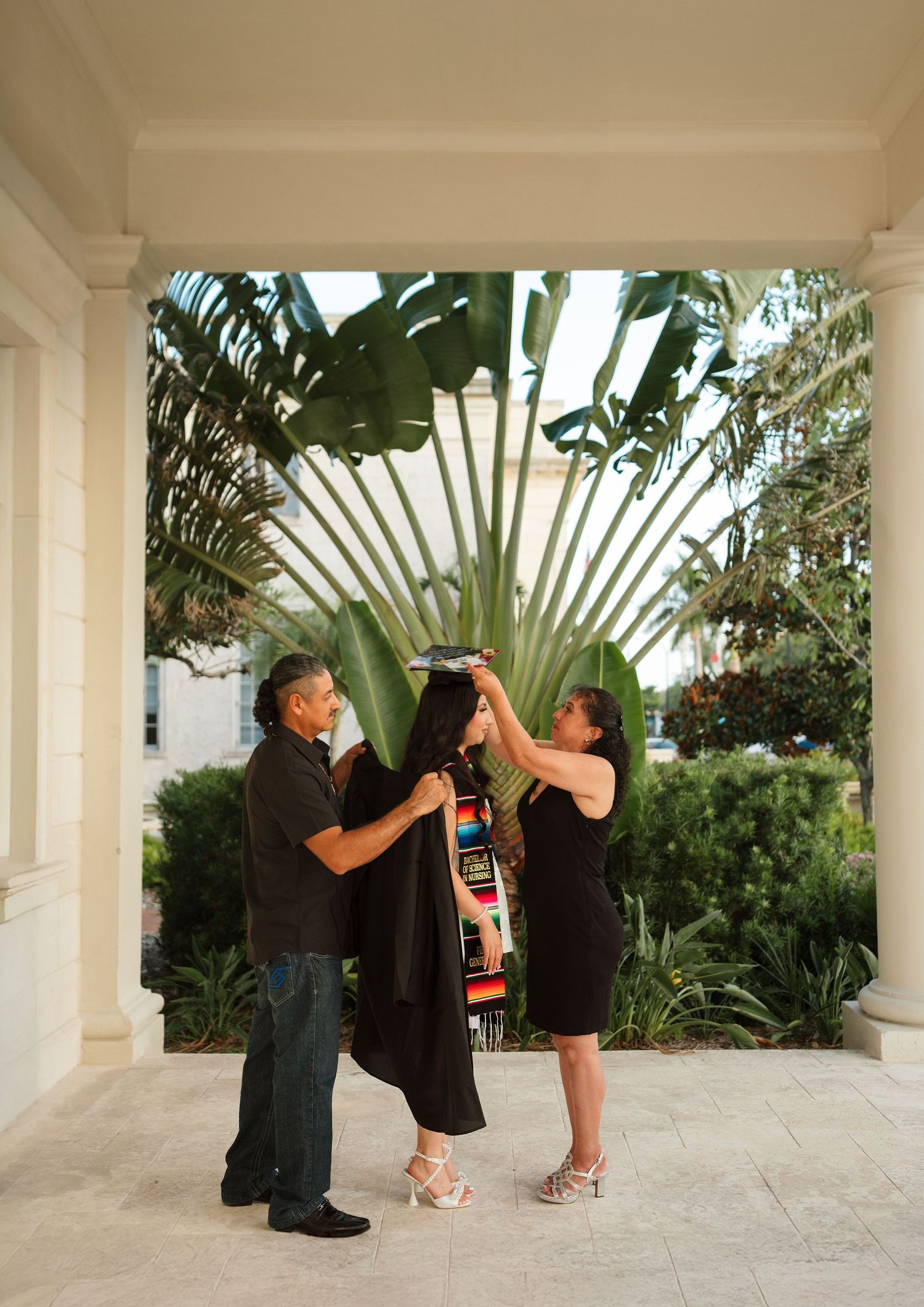 Parents helping graduate put on cap and gown outdoors under a columned portico with tropical plants.