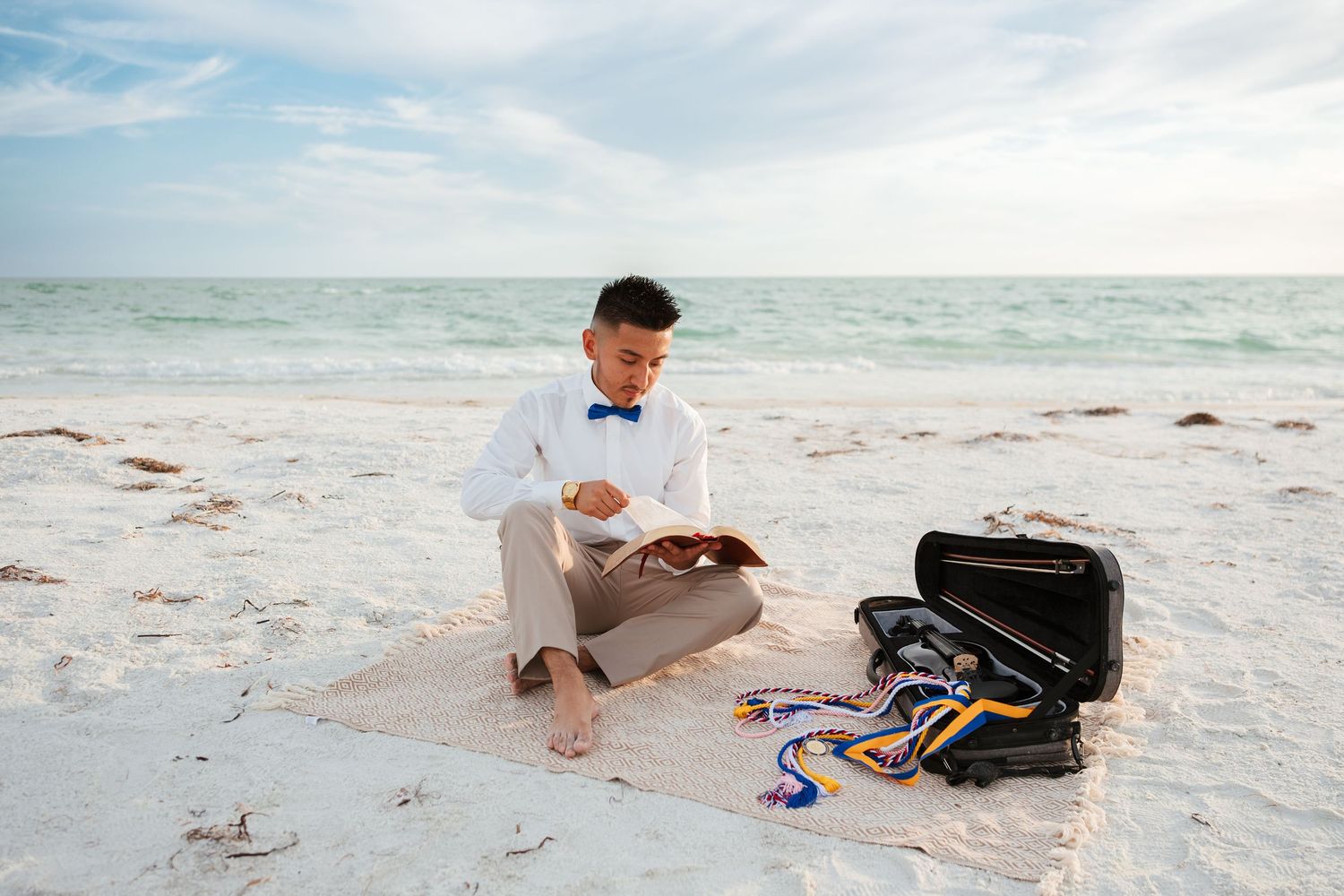 Young boy in bow tie suit reads book on beach blanket with open briefcase and medals beside him.