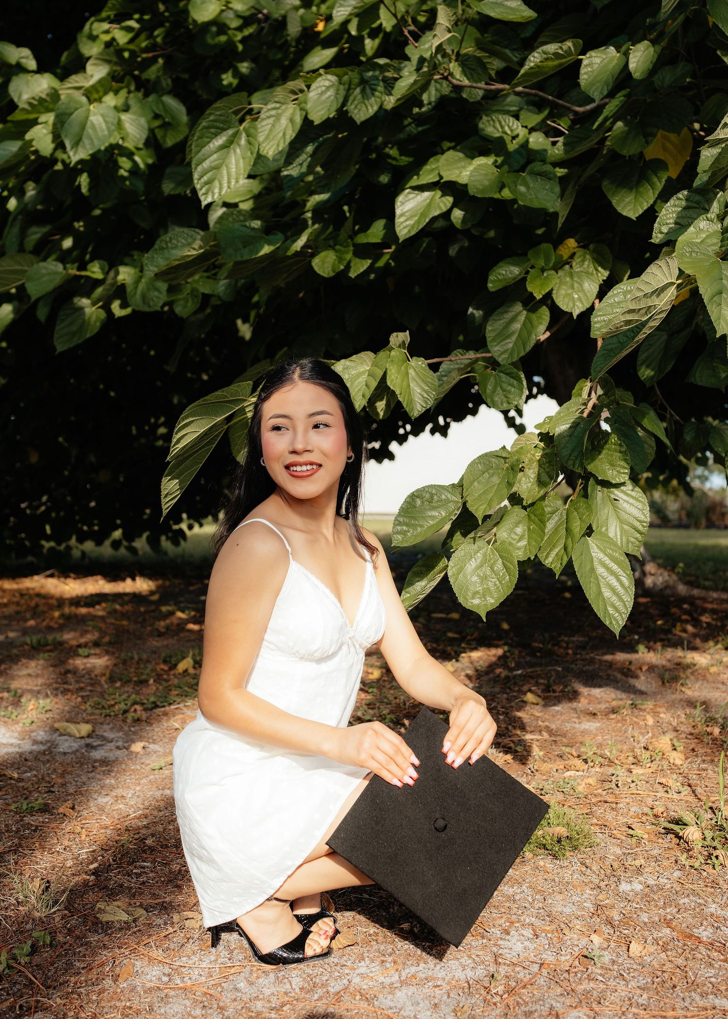 Young woman in white dress crouching outdoors holding graduation cap under green tree, smiling at camera.