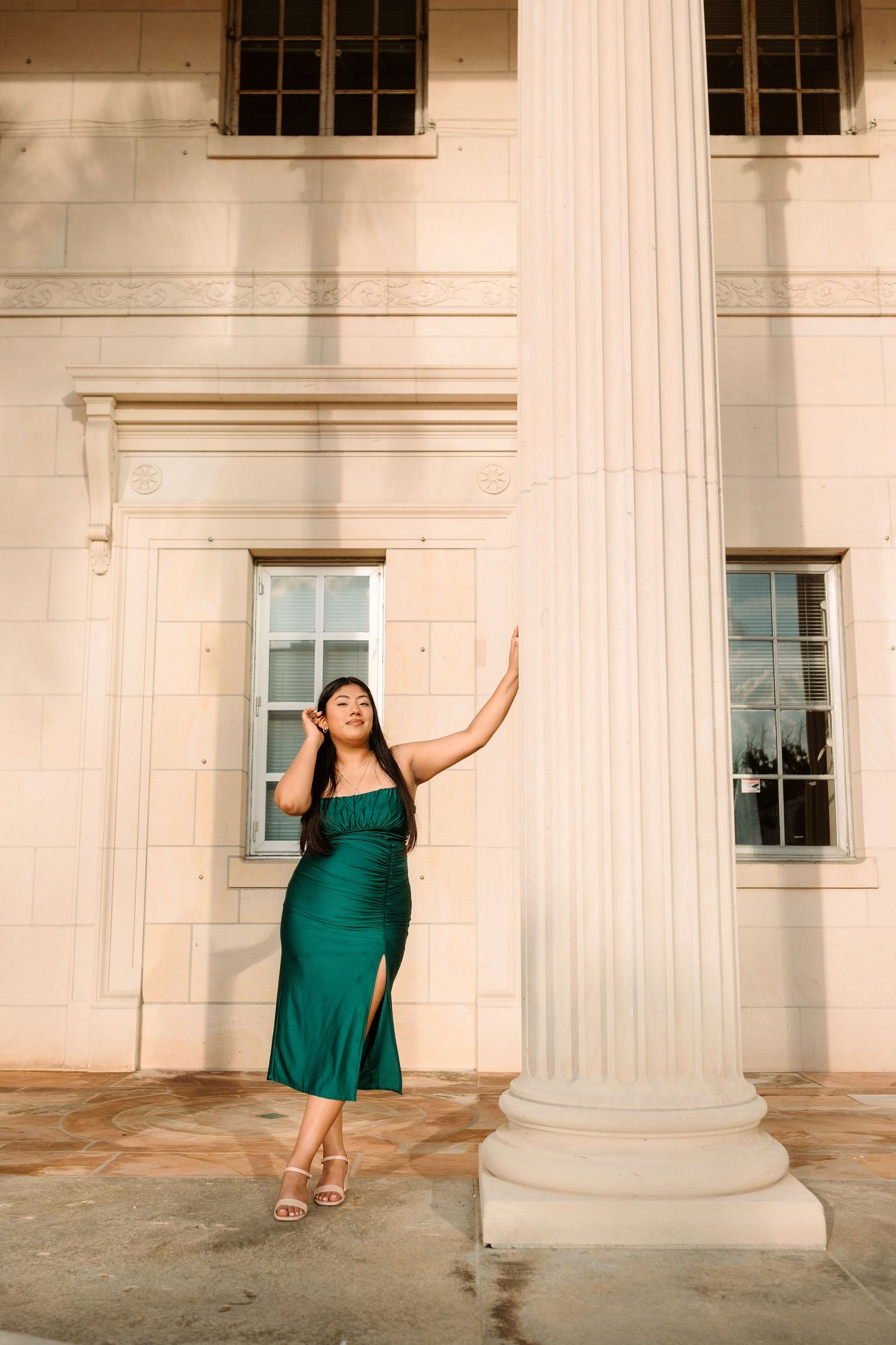 Woman in elegant green satin midi dress posing gracefully beside a white classical column outside a historic building.