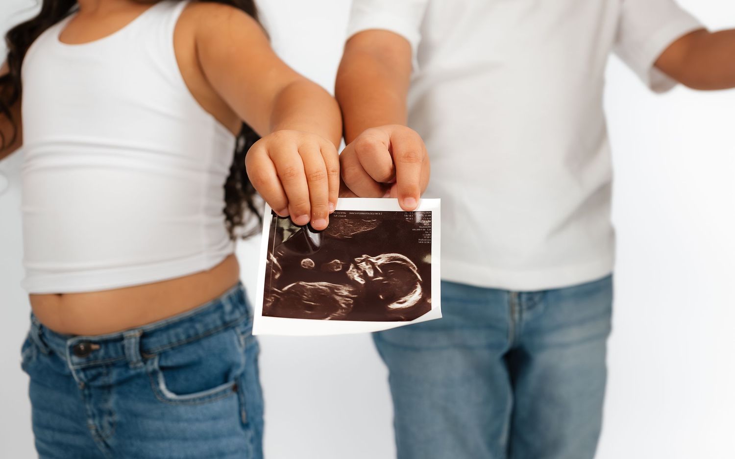 Two children in white shirts and jeans holding an ultrasound photo together against a white background.