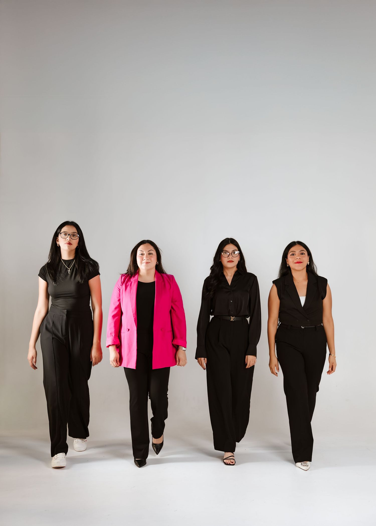 Four professional women in black outfits, one in pink blazer, walking confidently against a gray background.