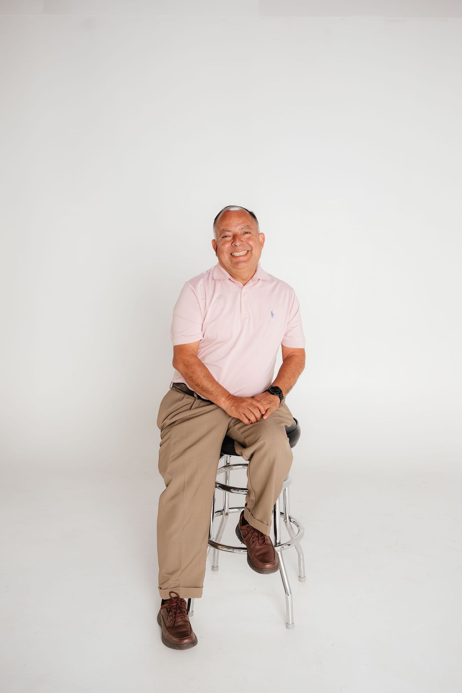 Middle-aged man in pink polo shirt and khaki pants sitting on stool, smiling against white background.