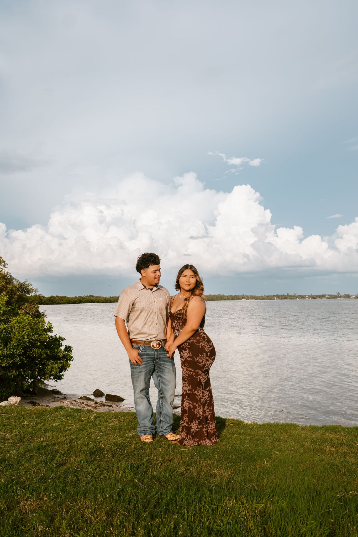 Couple holding hands standing on green grass by a scenic waterfront with dramatic white clouds in the background.