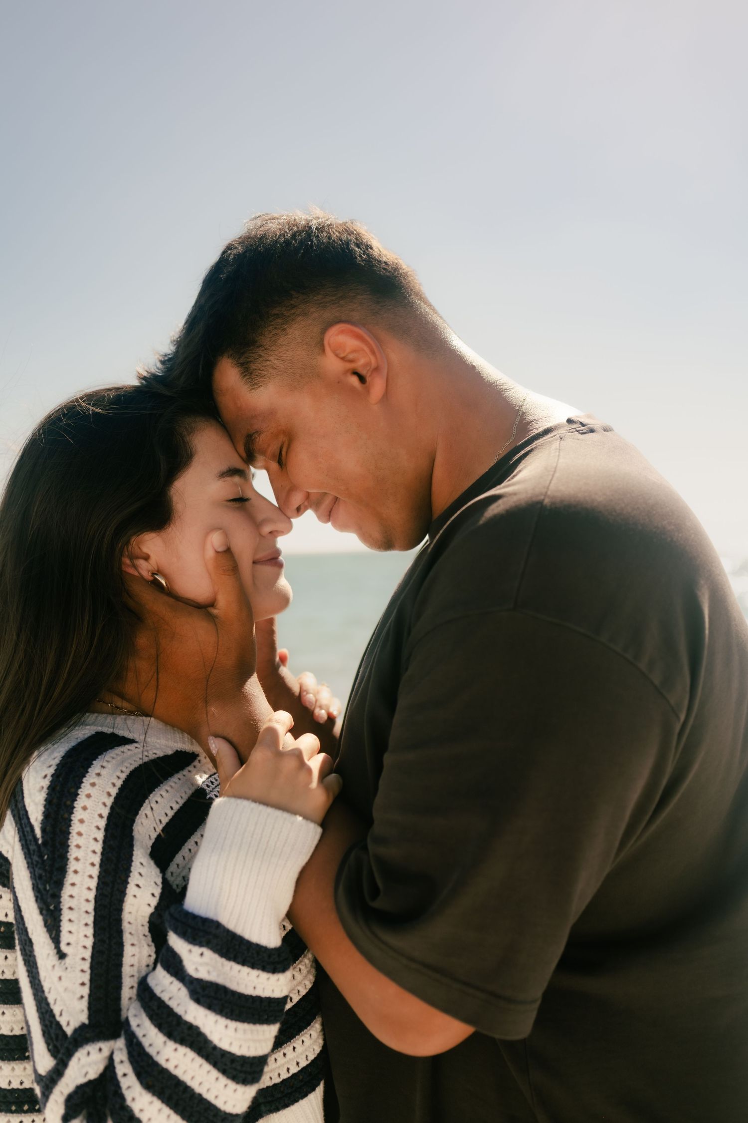 Romantic couple touching foreheads and smiling outdoors near the ocean on a sunny day.