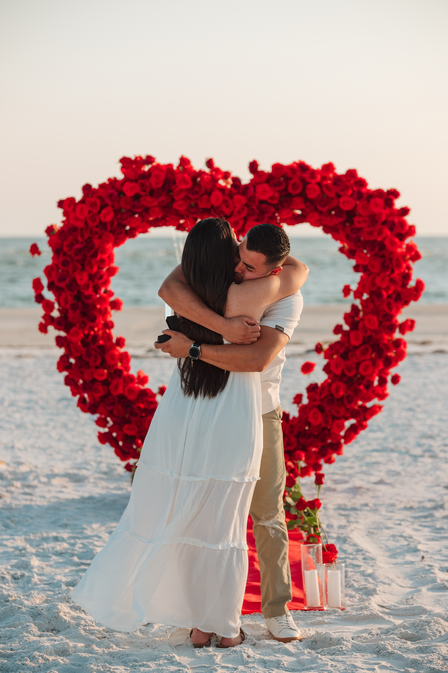 Couple embracing in front of a large red rose heart arch on a beach during a romantic proposal at sunset.