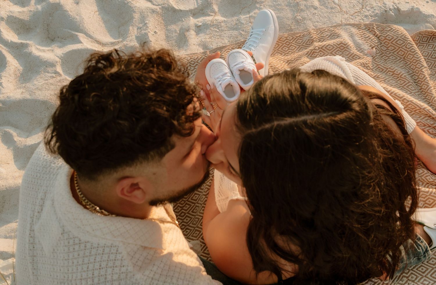 Couple lying on sandy beach sharing a romantic moment, woman wearing white sneakers, warm golden hour lighting.