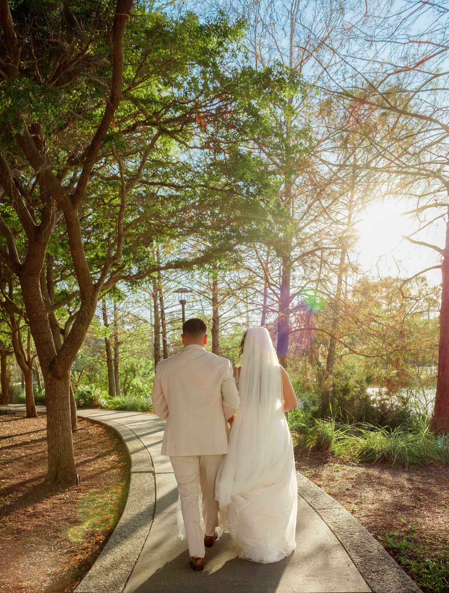 Bride and groom walking hand in hand along a sunlit garden path lined with trees.