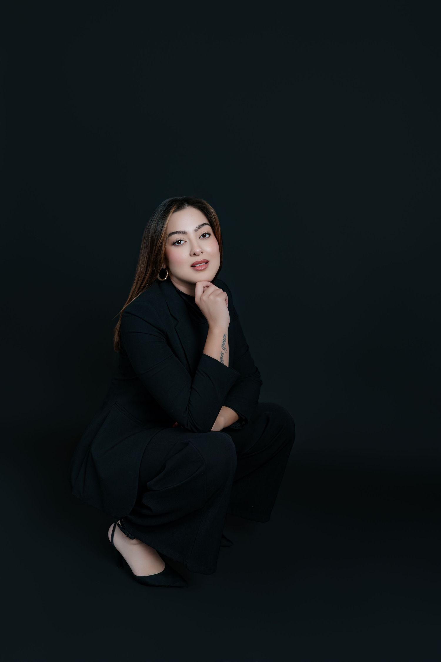 A young woman in a black suit sits gracefully against a dark background, resting her chin on her hand.