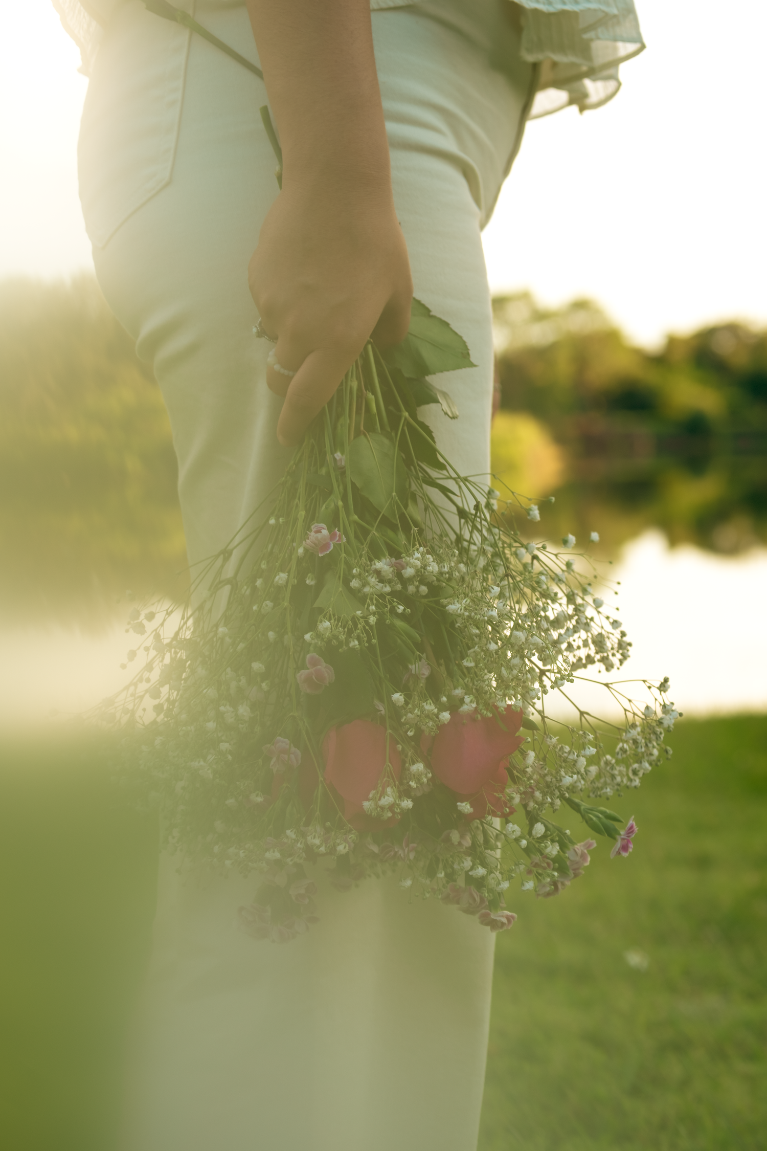 Person in white pants holds a delicate bouquet of wildflowers against a sunlit outdoor background.
