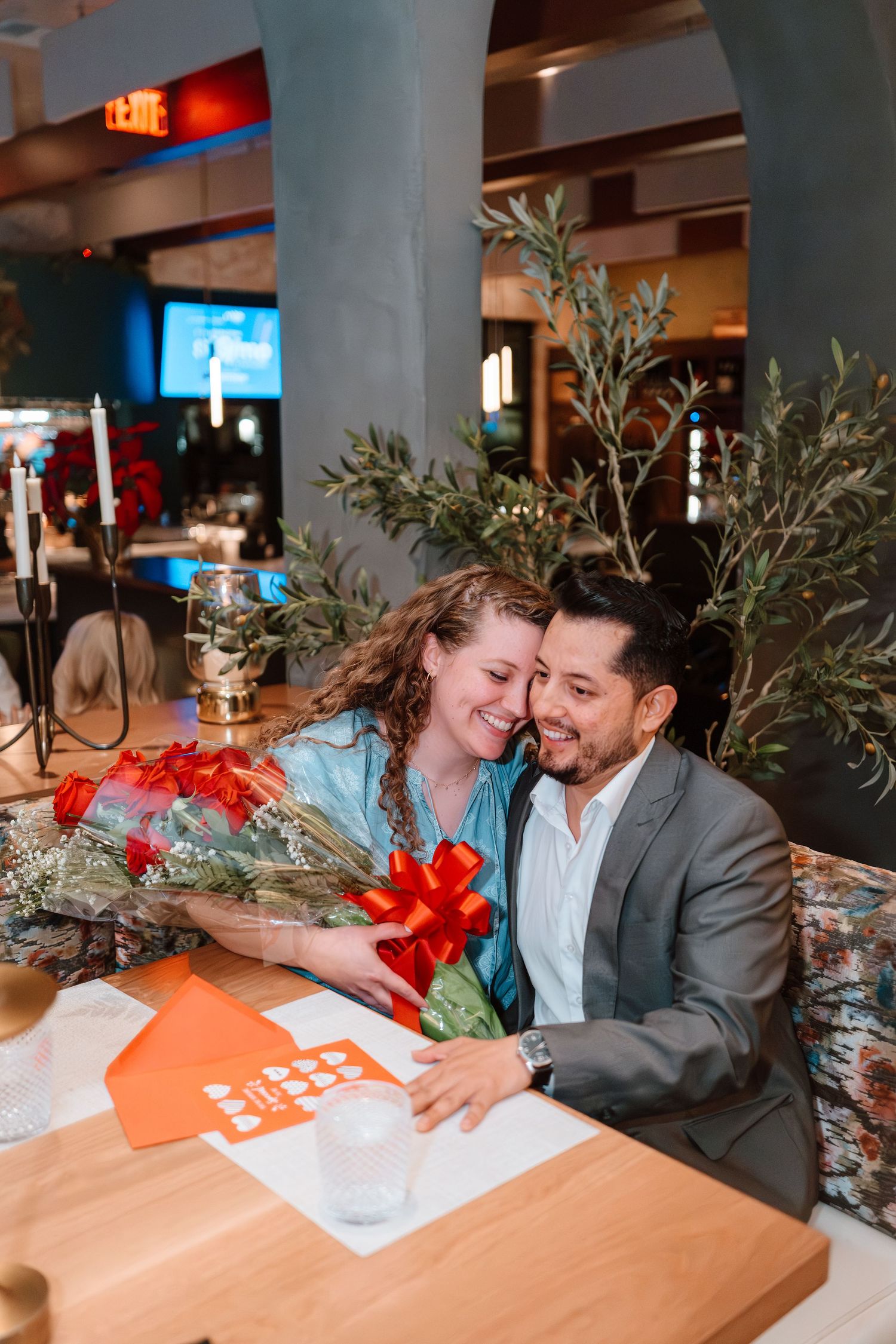 Couple embracing at restaurant table with red bouquet and gift, smiling joyfully in elegant dining setting.