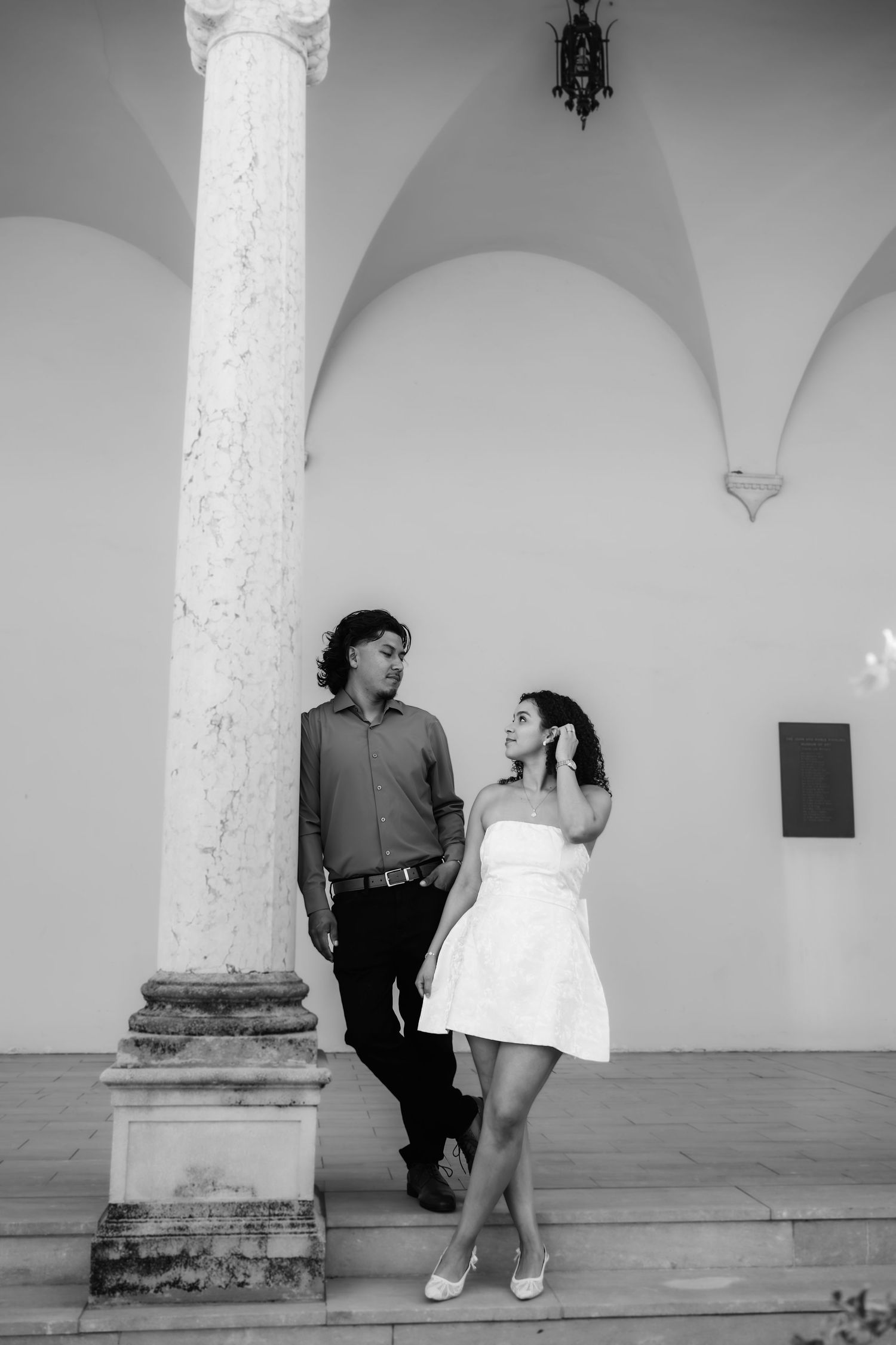 Black and white photo of a couple standing by a classical column under arched architecture, sharing a romantic moment.