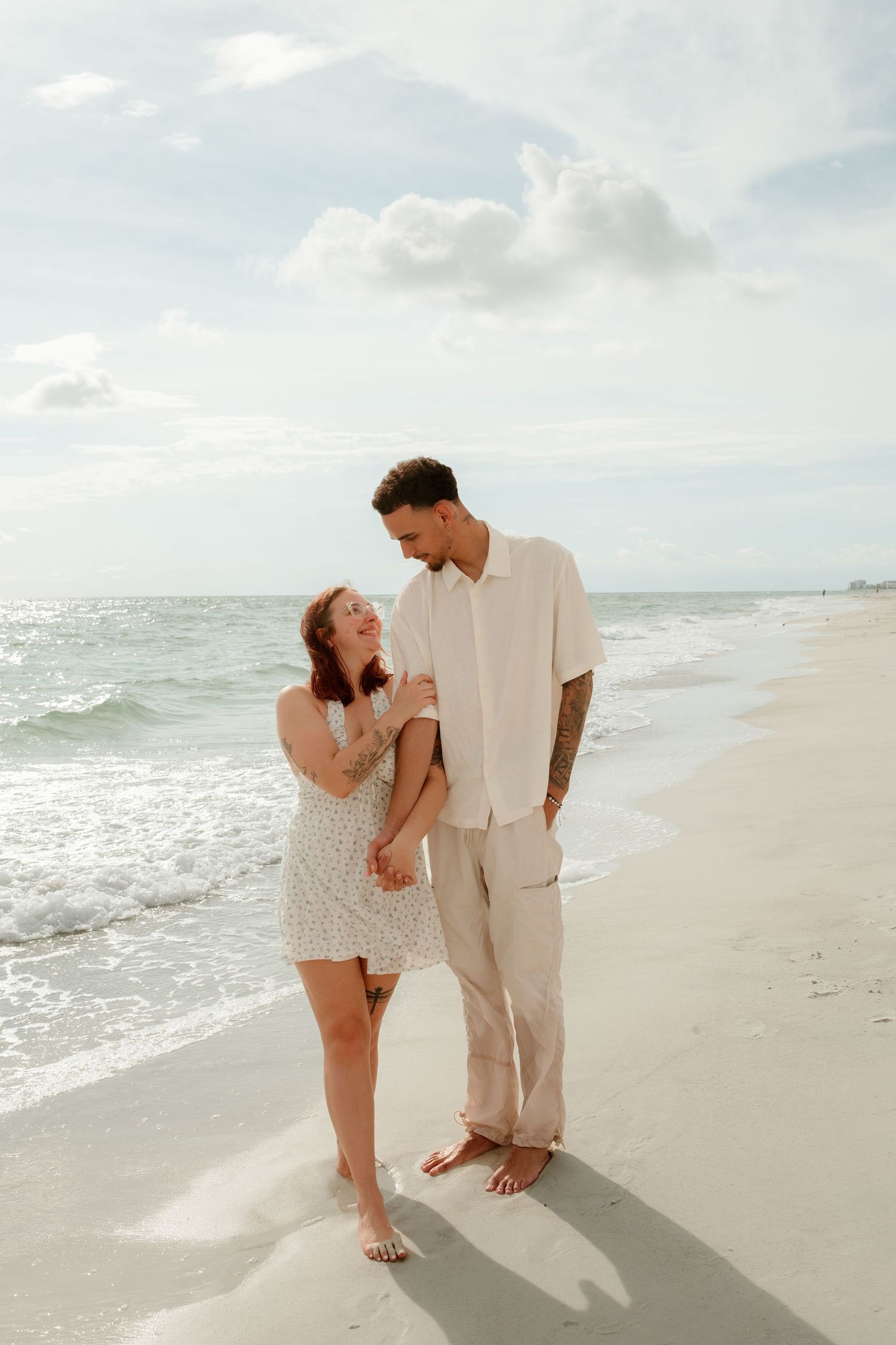 A couple walks barefoot on a sandy beach, smiling at each other in light summer outfits with ocean waves behind them.