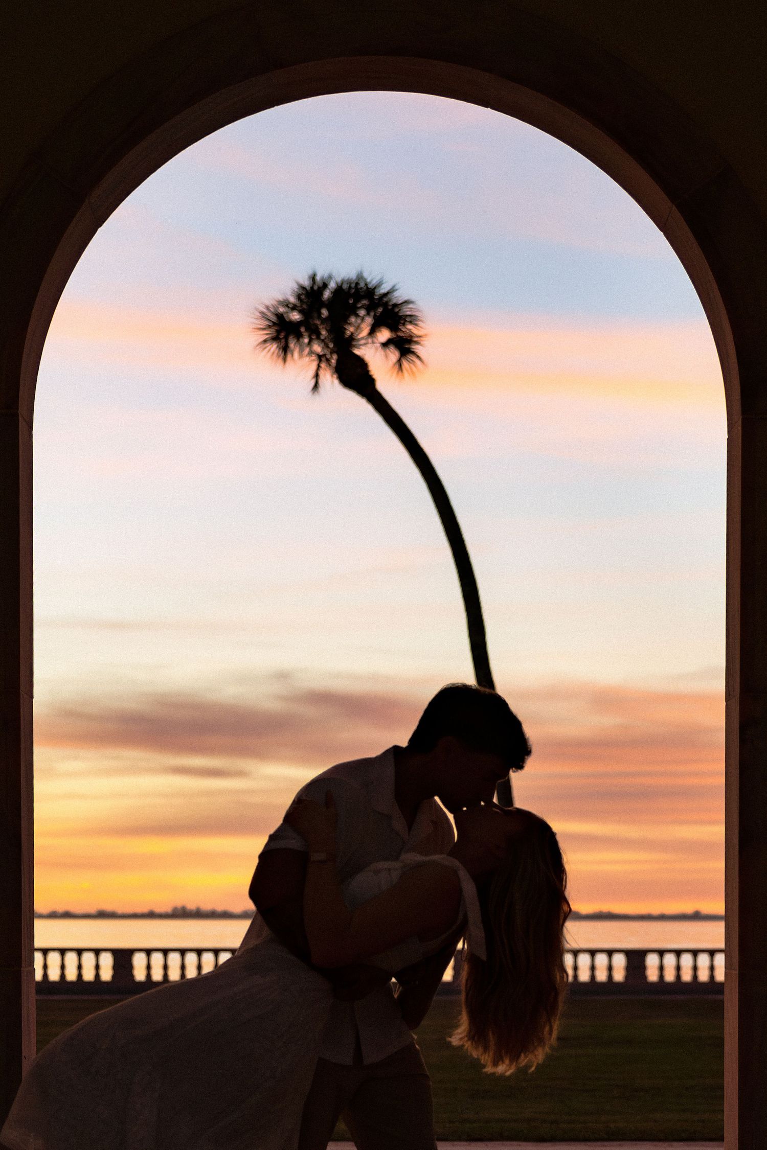 Romantic silhouette of couple kissing under an arch at sunset with a palm tree and colorful sky.