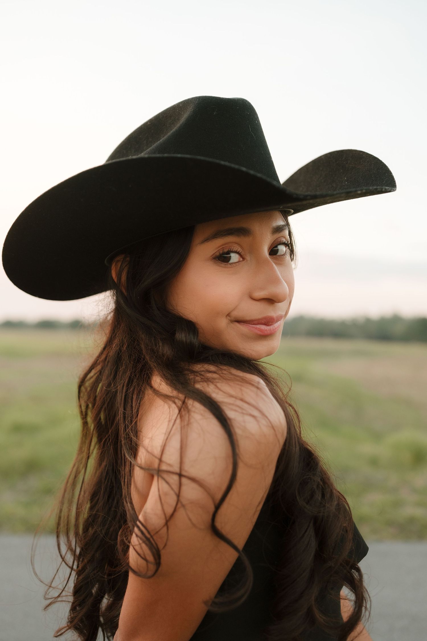 Young woman in black cowboy hat with long dark hair smiling over shoulder outdoors at dusk.