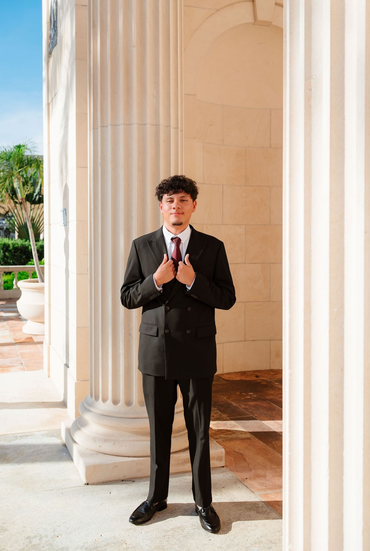 Young man in black suit adjusts tie while standing between white classical columns of elegant building.