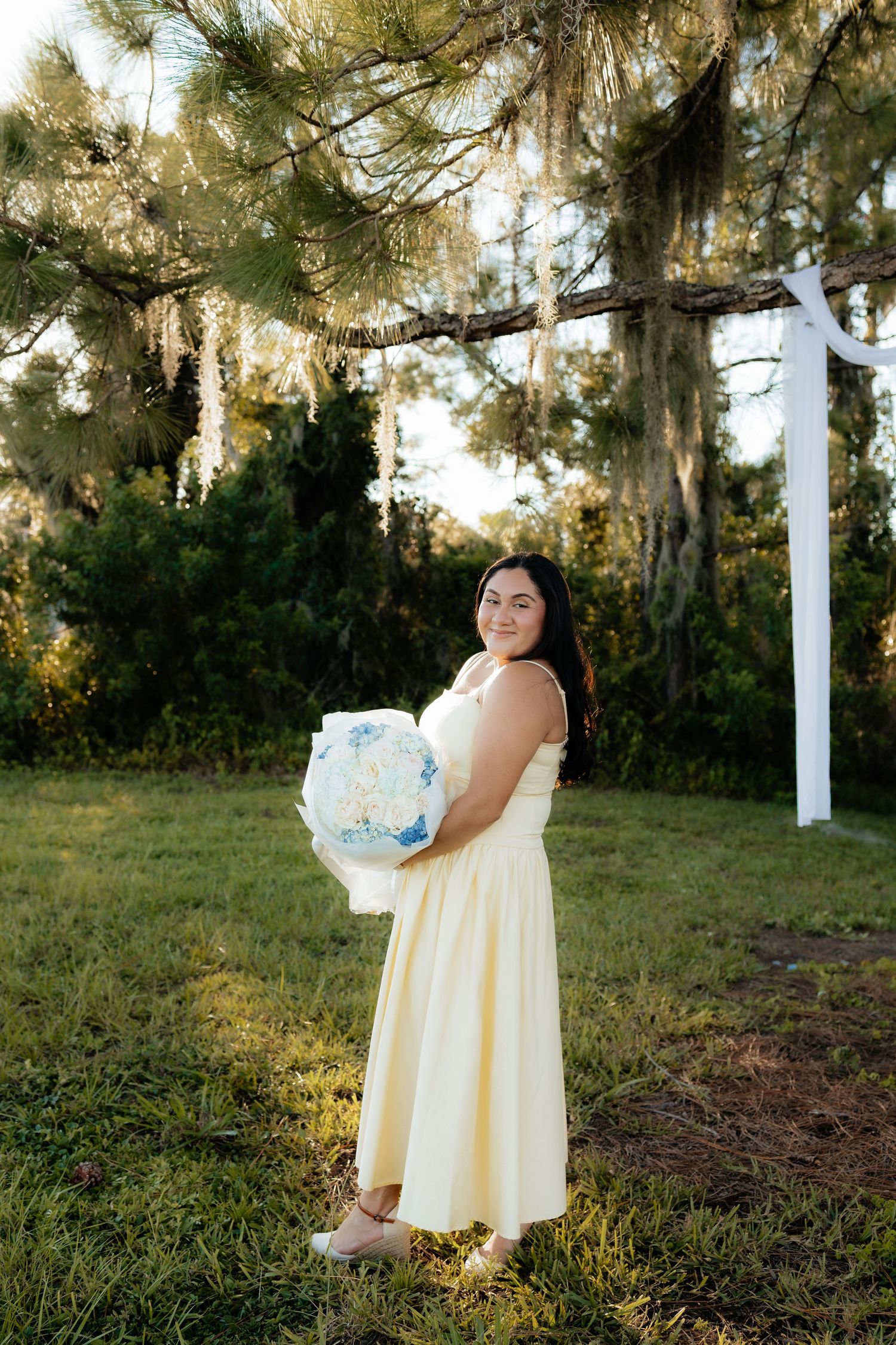 Woman in yellow dress holding blue floral bouquet outdoors near pine trees with Spanish moss at golden hour.