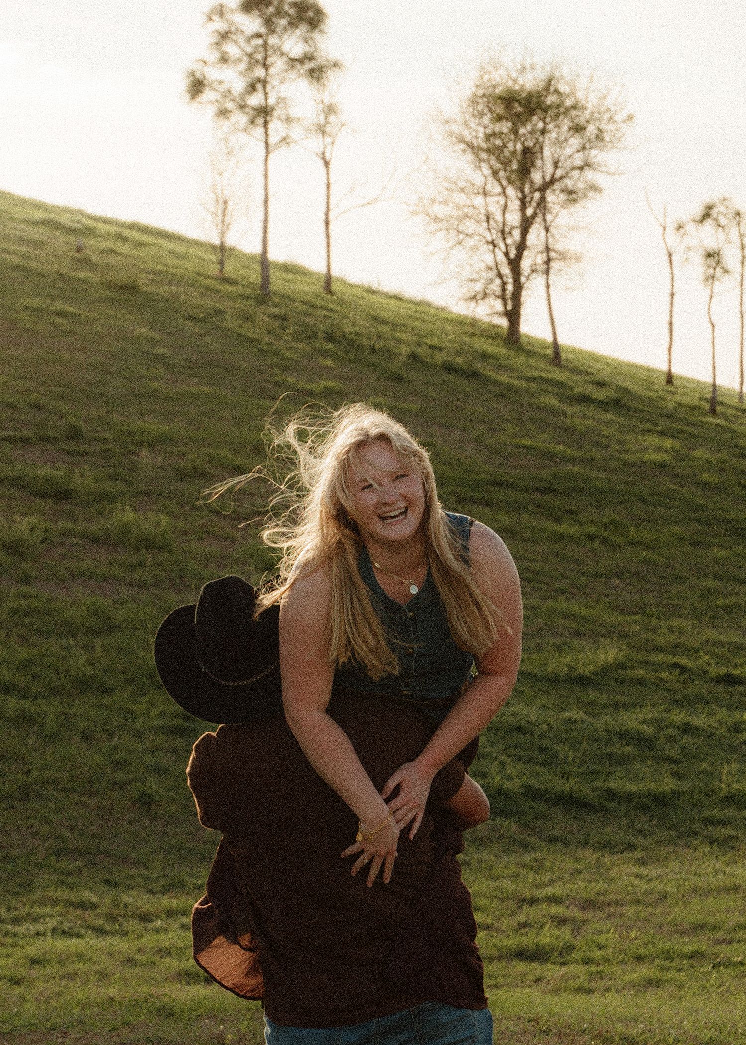 A laughing blonde woman being lifted by a person in a cowboy hat on a sunny green hillside with trees.