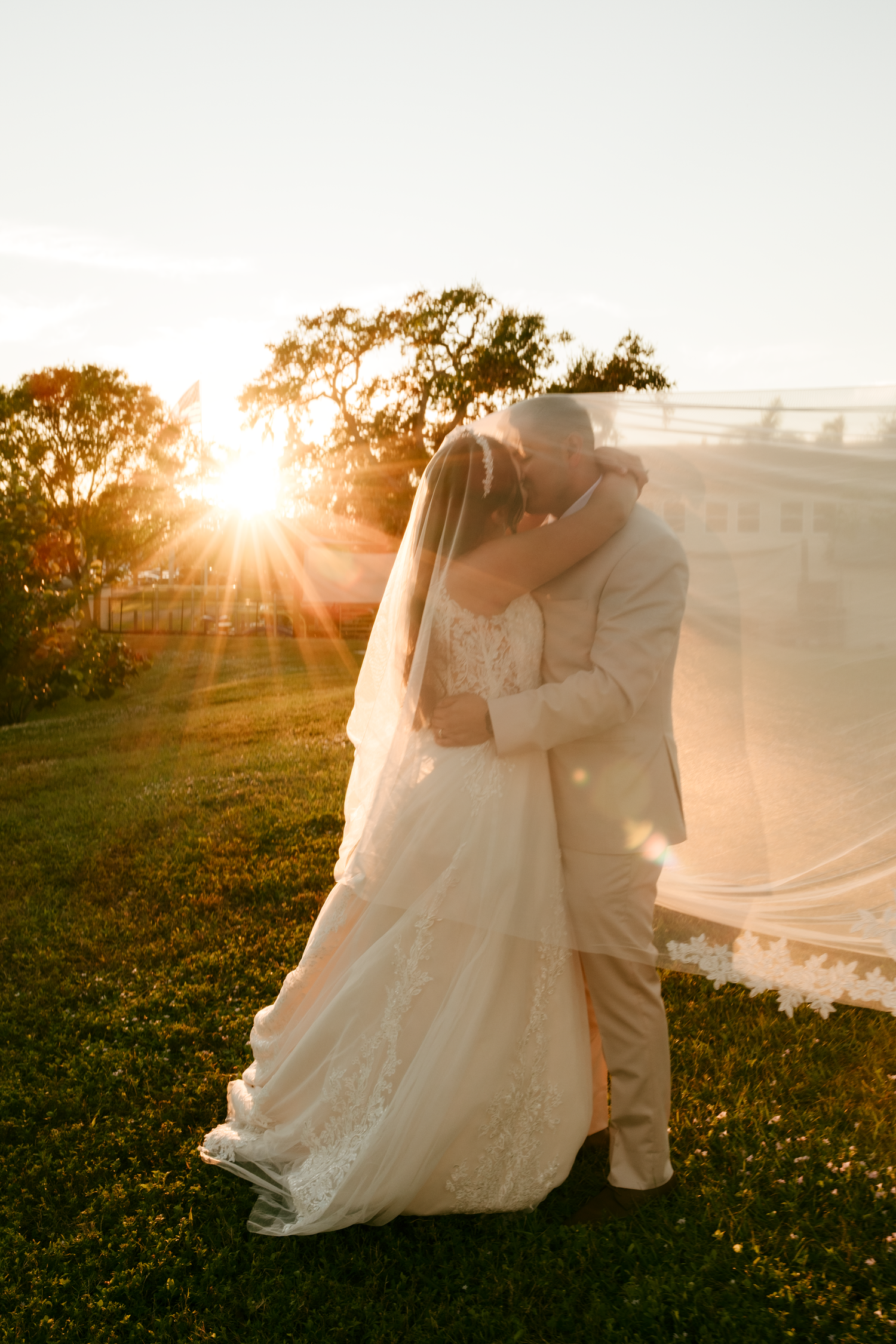 Bride and groom share romantic sunset kiss outdoors with golden light and flowing veil.