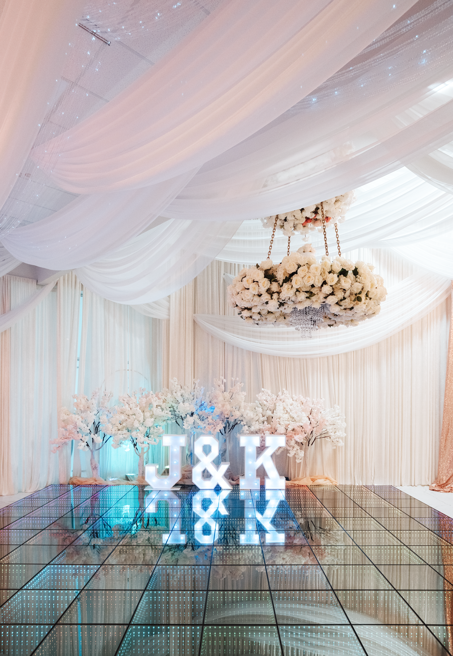 Elegant wedding reception venue with J&K marquee letters, mirrored dance floor, and floral chandelier.