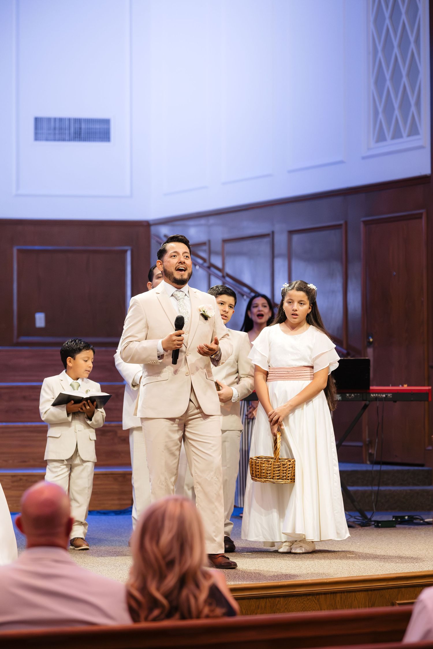 Groom in tan suit speaks at church altar flanked by flower girl and ring bearer during wedding.