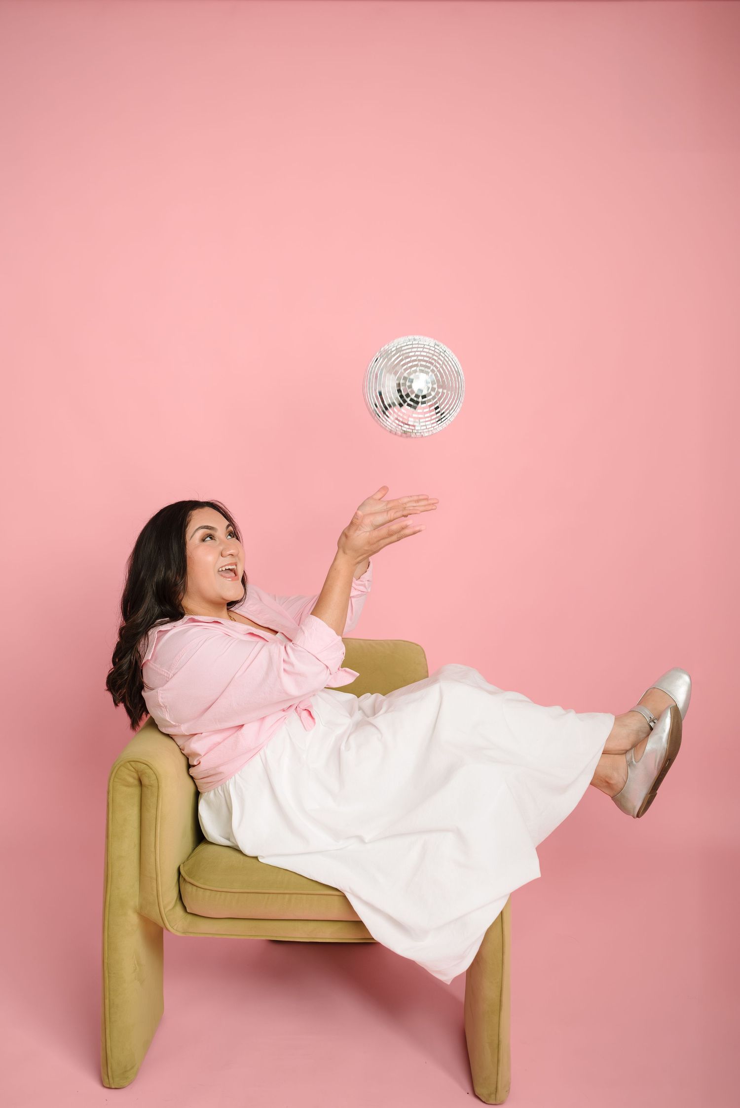 Woman in pink blouse and white skirt sitting in chair tossing a disco ball against pink background.